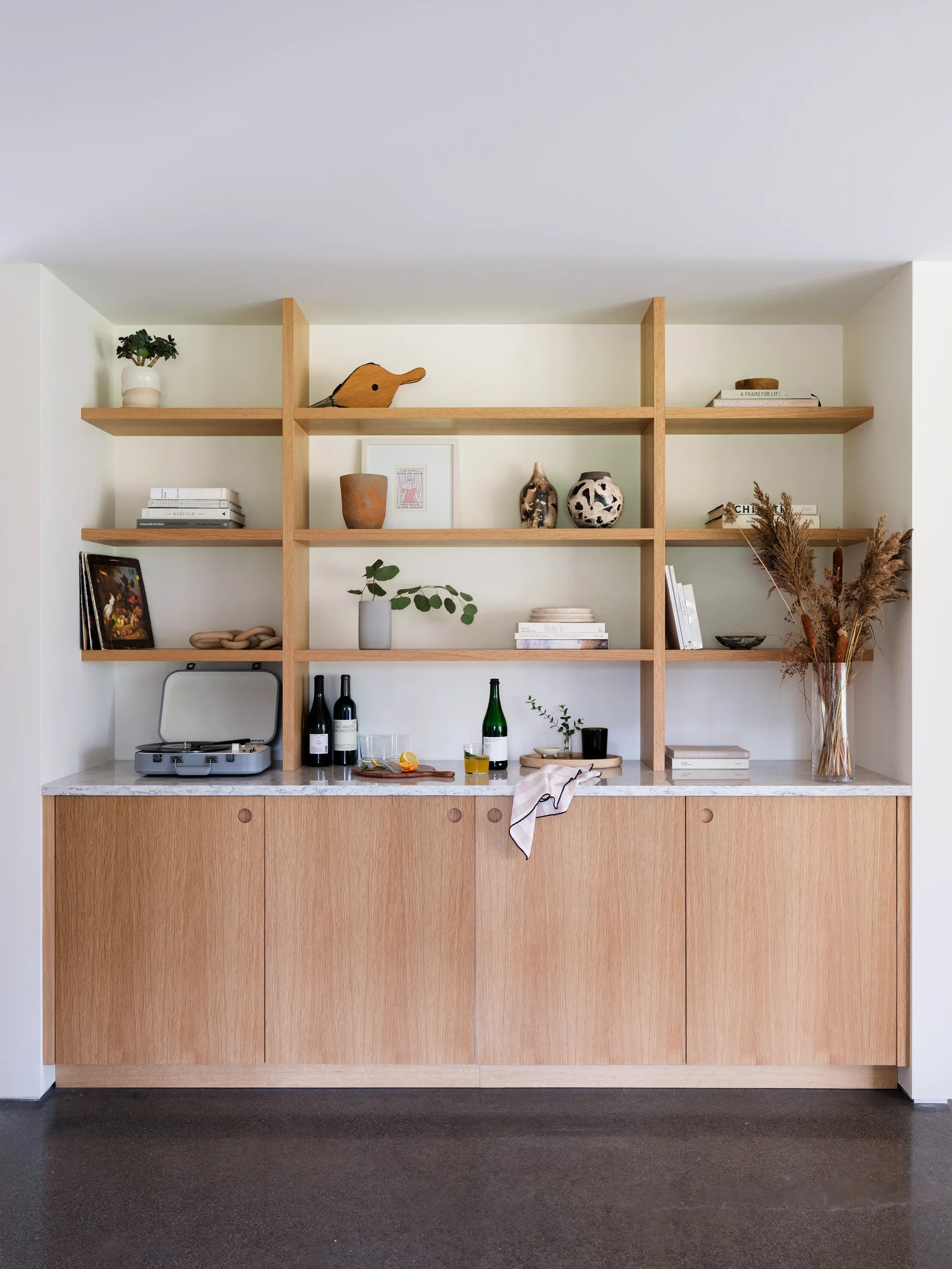 A wooden cabinet with a white marble top, on which sits a gramophone, three bottles, and a glass of drink. The cabinet has open shelves with books, artwork, pottery, plants, and dried flowers arranged on them.