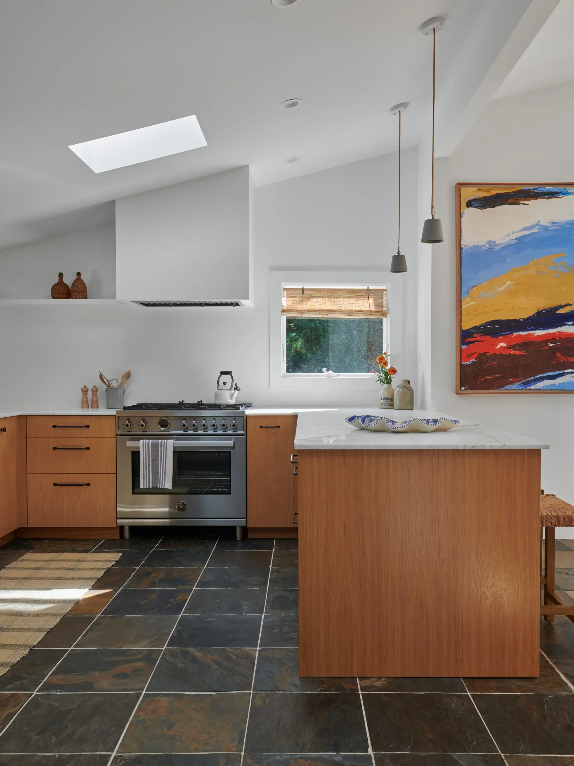 Interior of a modern kitchen with a white ceiling, skylight, and hanging pendant lights. The kitchen features a wooden island with a marble countertop, a stainless steel oven, and a window with a bamboo shade. There are decorative vases and a colorful abstract painting on the wall.