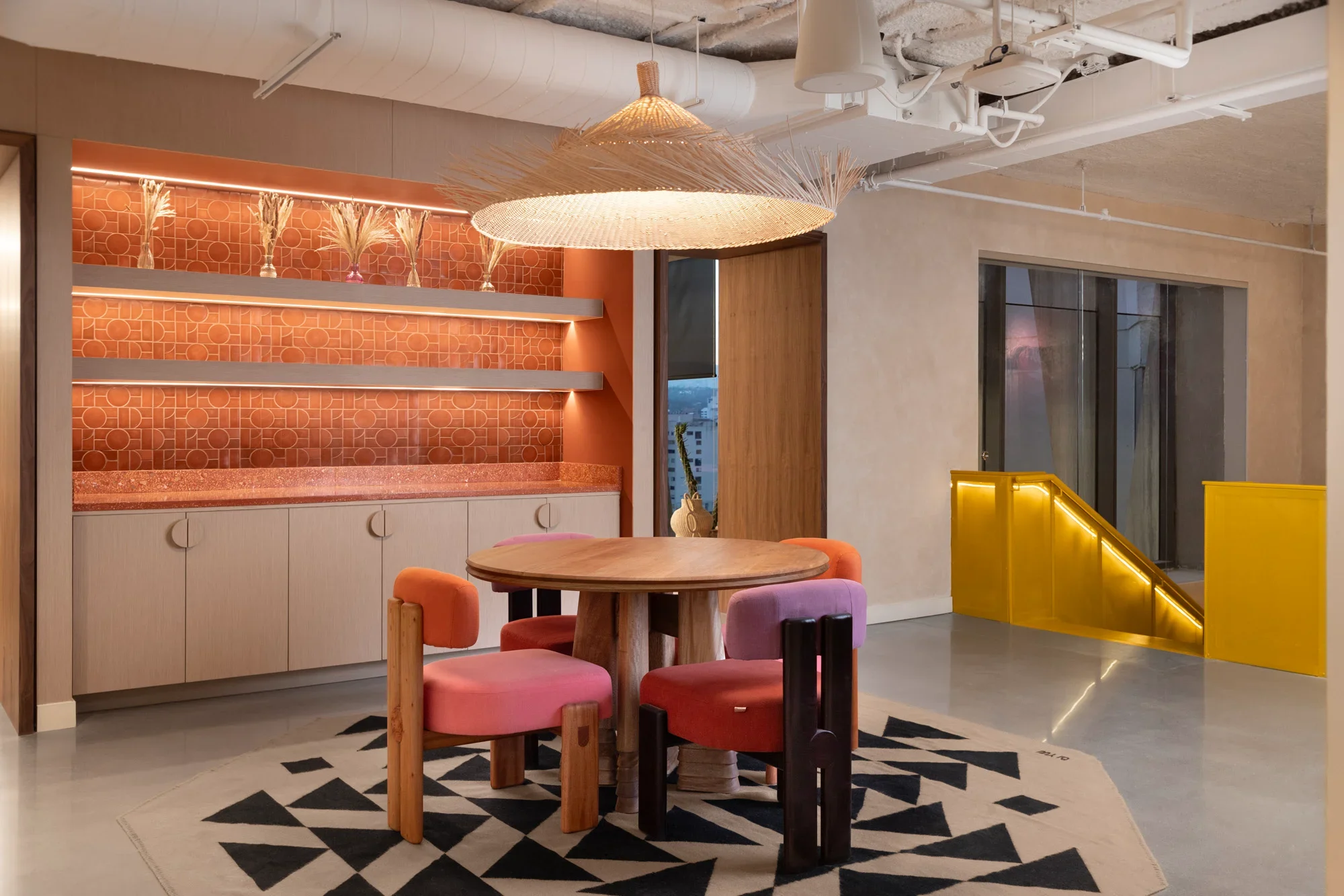 Interior of a modern room with a round wooden table surrounded by pink upholstered chairs, a black and white geometric rug, a built-in cabinet with orange vases and pampas grass, a large woven pendant light, yellow stairs with LED lighting, and large windows showing an urban view.