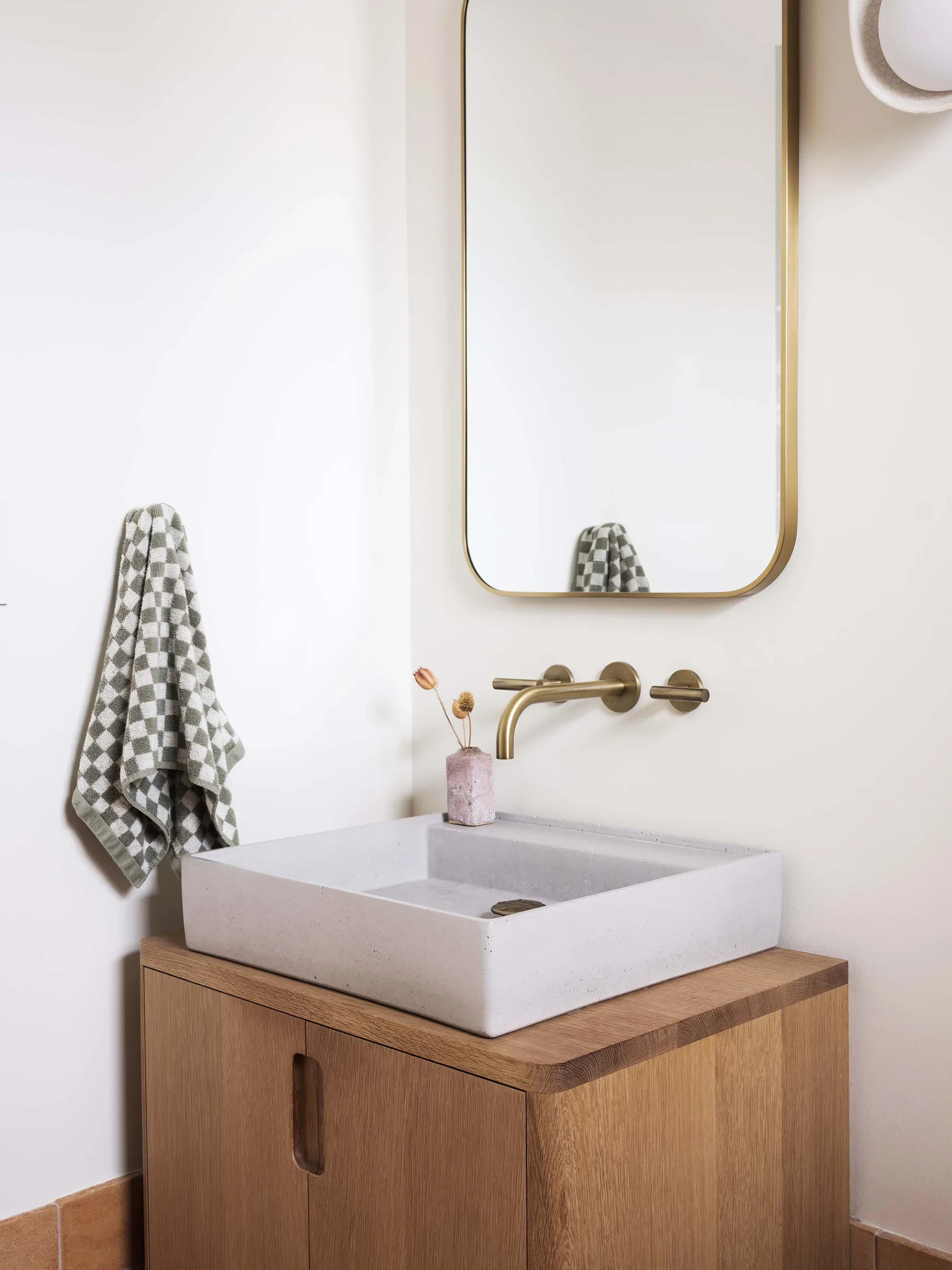 A modern bathroom vanity with a square white sink, a gold faucet, a wooden cabinet, a wall mirror, and a patterned towel hanging on a hook
