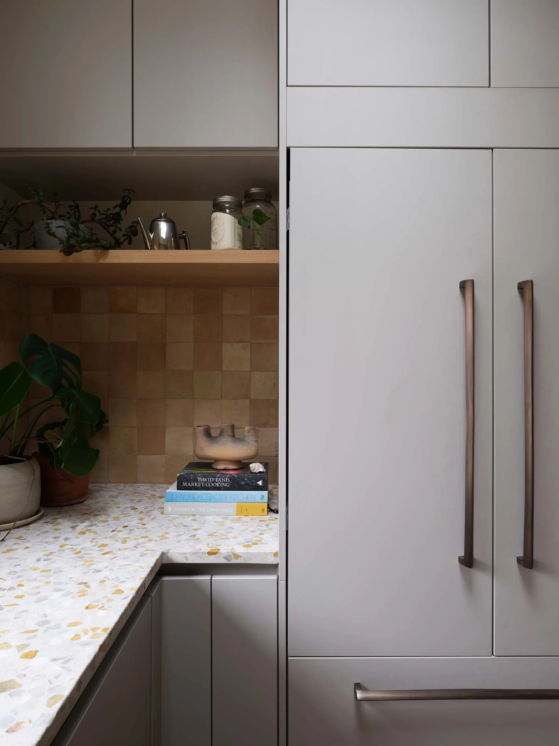 Kitchen with gray cabinets, a terrazzo countertop, and a backsplash with beige and brown tiles. There are potted plants, glass jars, and a book on the counter. The visible book titles are "Market Cooking," "My Mexico City Kitchen," and "Dinner at the Long Table." The right side shows a closed gray cabinet with long, curved wooden handles.