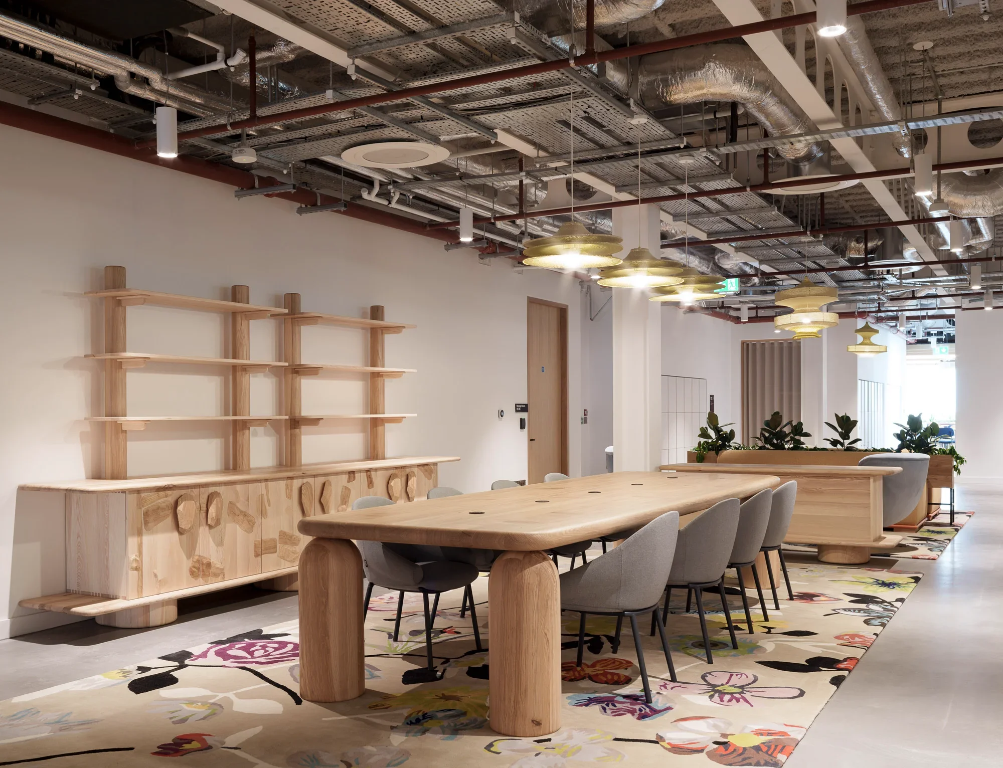 Modern office interior with a large wooden conference table surrounded by gray chairs. The ceiling features exposed ducts, pipes, and lighting fixtures.