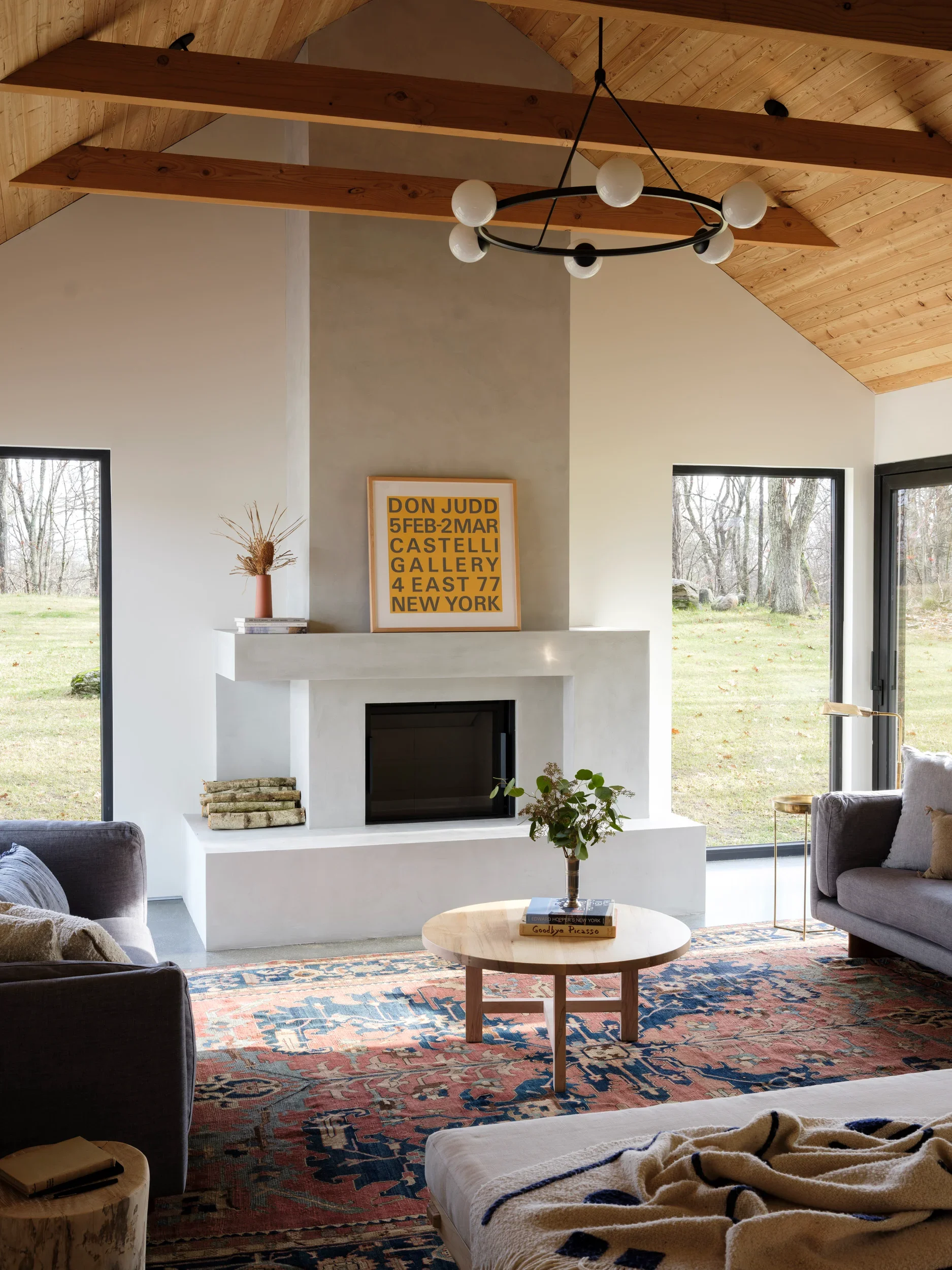 A cozy living room with a fireplace, surrounded by large windows showing a backyard with trees. There is a colorful patterned rug on the floor, a round wooden coffee table with books on it, and multiple seating options including a beige armchair with blankets and a small side table.