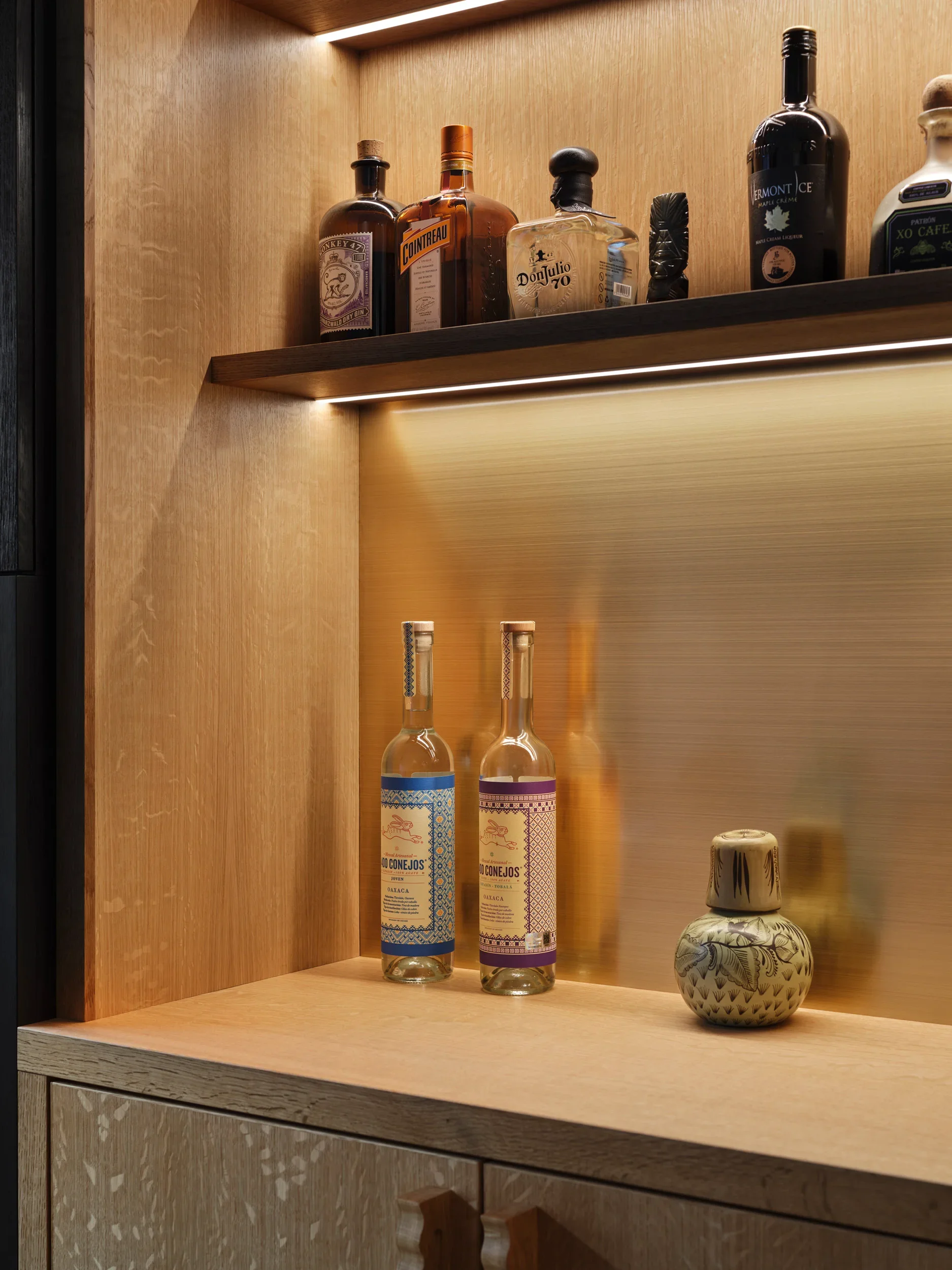 Wooden cabinet with a bottle of Cointreau, Don Julio tequila, a black liquor bottle, and a ceramic decorative bottle inside the lit shelves.