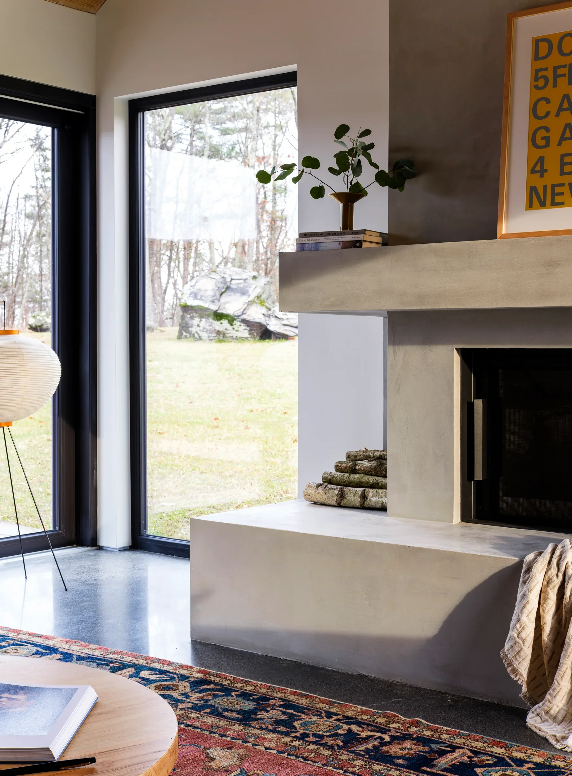 Interior of a modern living room with a fireplace, a windows with black frames showing an outdoor view of trees, a round wooden table with an open book, a patterned rug, and a corner with logs and a framed artwork.
