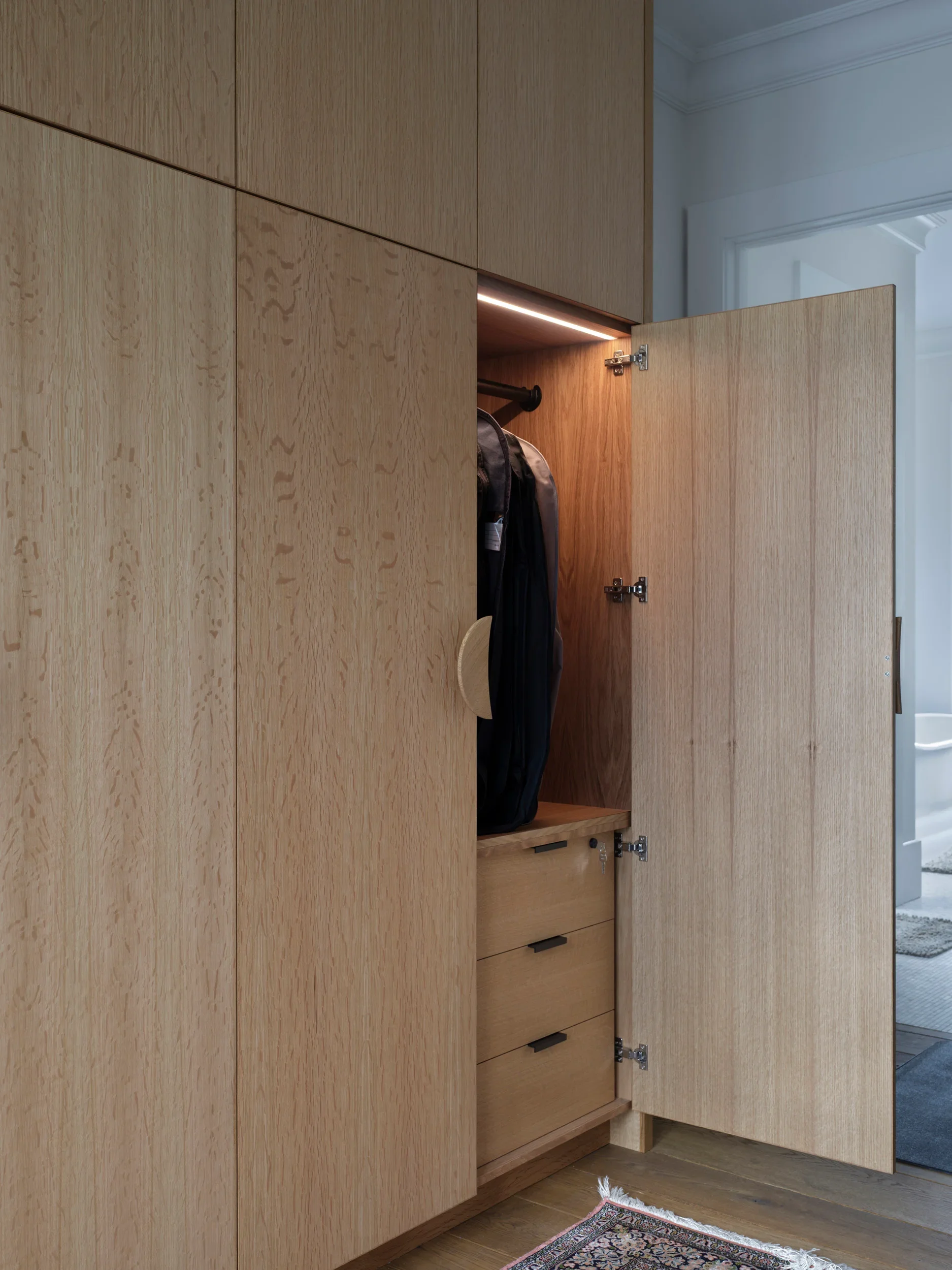 Open wooden cabinet with clothes hanging inside and cabin detailing, located in a room with a patterned rug and wooden flooring.