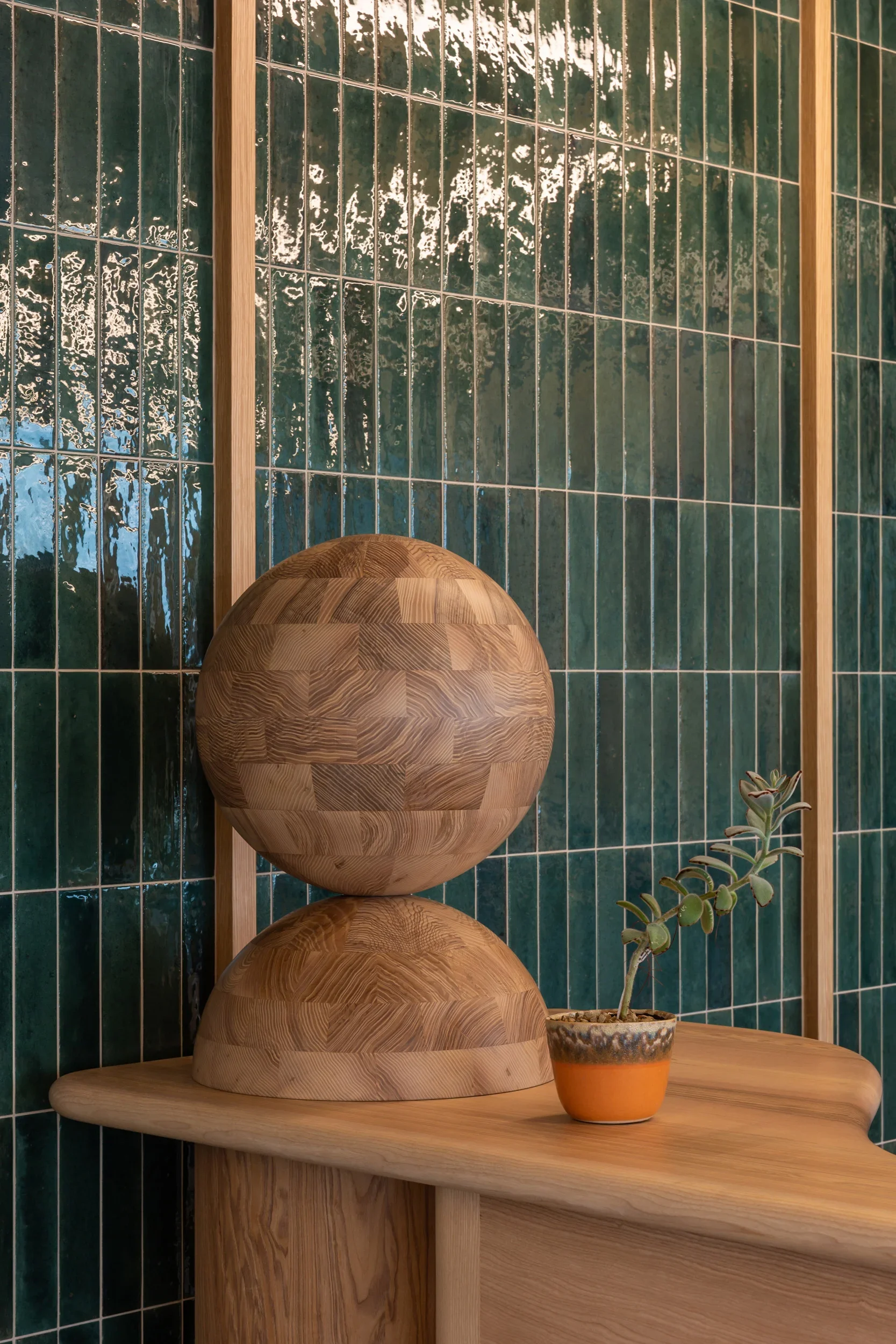 Three wooden spheres stacked on top of each other on a wooden shelf, with a small potted succulent plant to the right, against a background of green tiled wall with white grout lines.