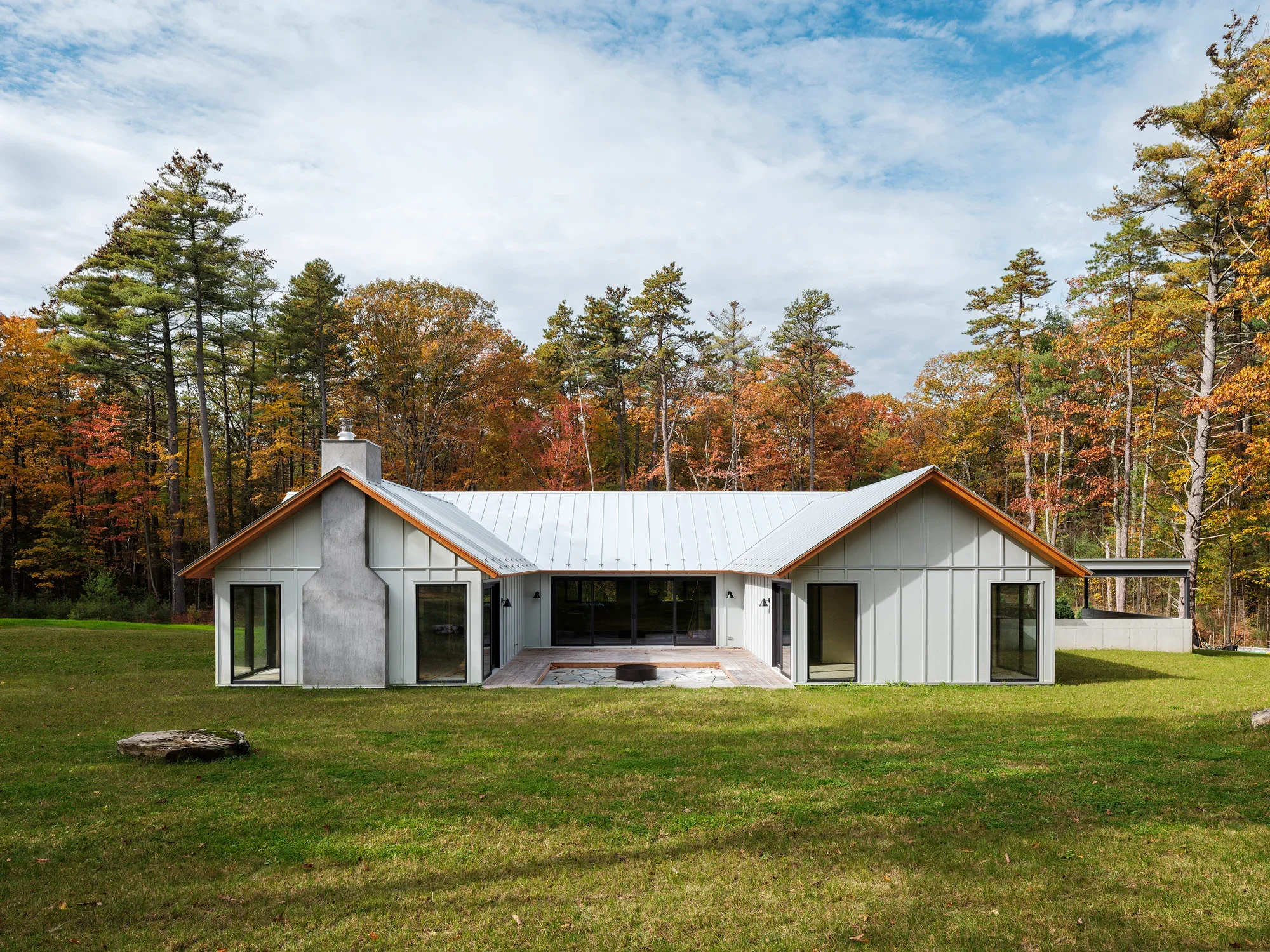 A modern house with large glass doors, surrounded by a well-maintained lawn and a backdrop of tall trees with colorful fall foliage under a partly cloudy sky.