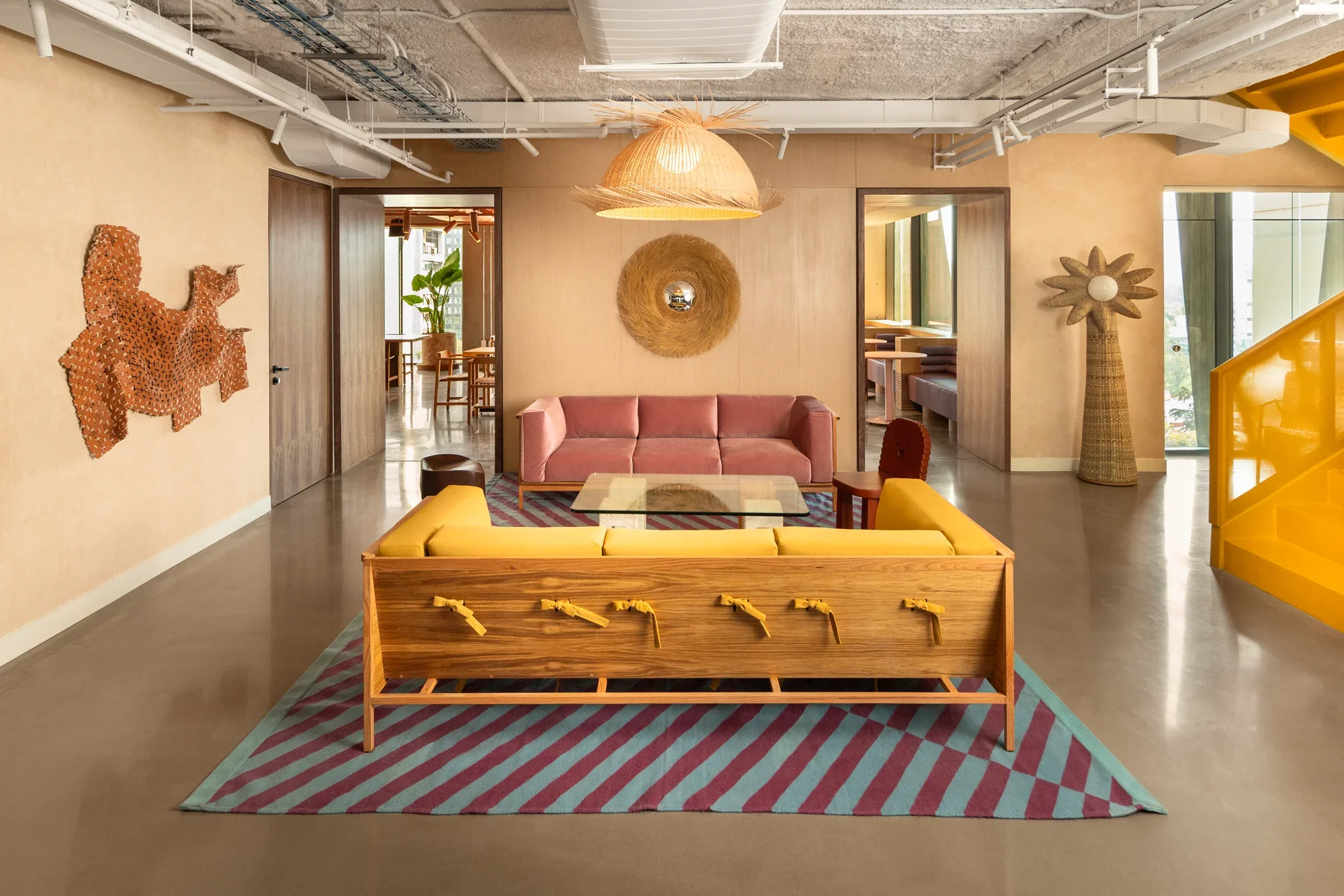 Interior of a modern office space with a pink sofa, a hanging bamboo light fixture, decorative wall art, and a yellow staircase on the right side. There is a colorful striped rug under a wooden table with yellow ties, and large windows on the right allowing natural light.