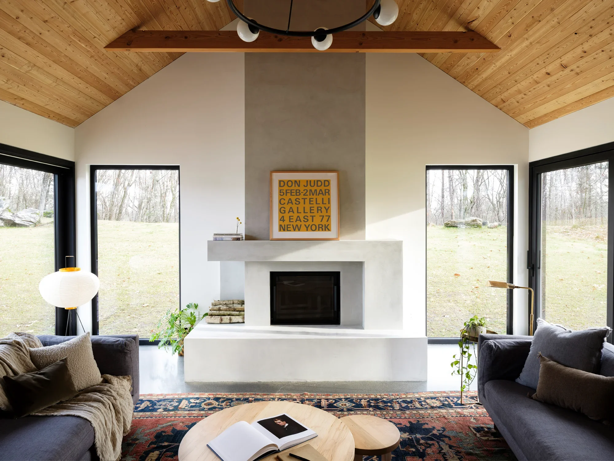 Living room with a white fireplace, windows with a view of trees outside, a black sofa on the right, a beige sofa on the left, a wooden coffee table with an open book, plants, a colorful rug, and a wooden ceiling with a black spherical chandelier with four light bulbs.