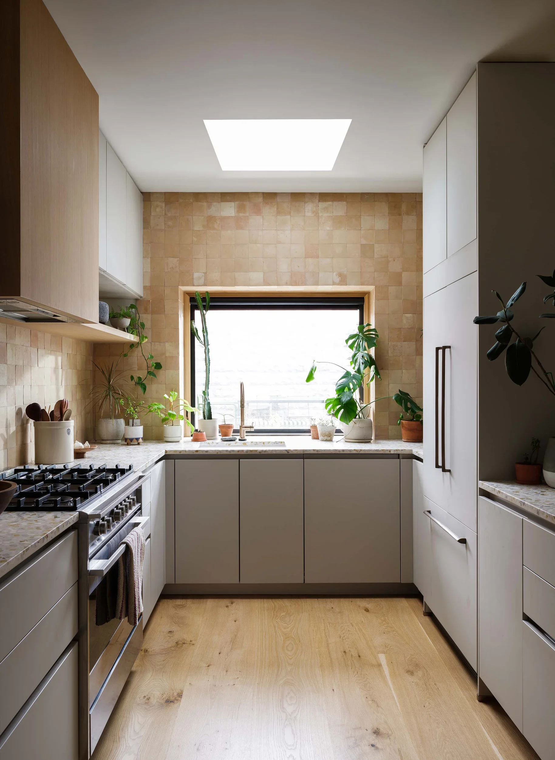 Modern kitchen with wood and white cabinets, beige tiled wall, large window with plants, granite countertops, and stainless steel appliances.