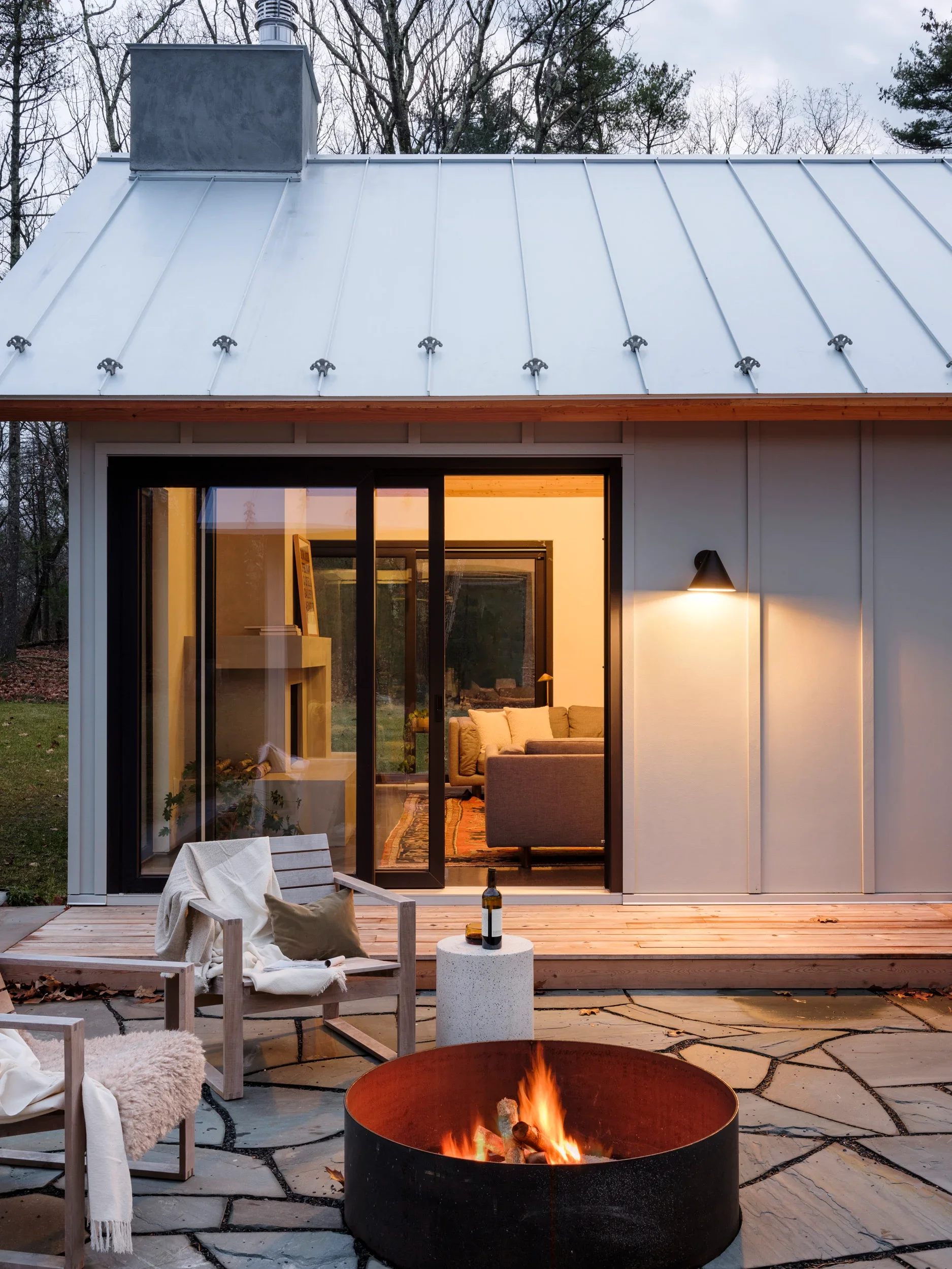 A cozy outdoor patio with a fire pit, lounge chairs, and a small table, connected by sliding glass doors to a warm living room inside. The house features a metal roof and is surrounded by trees.