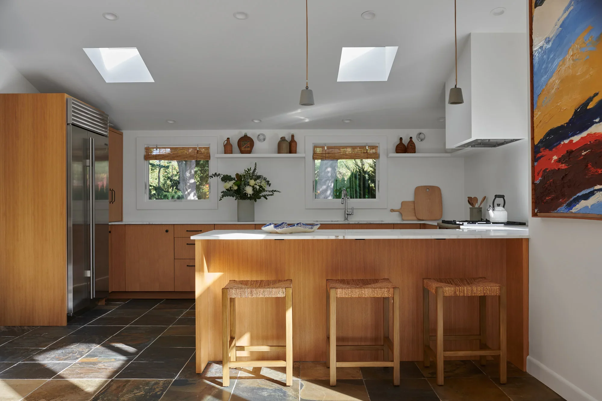 Modern kitchen with wooden cabinets, a white countertop island, and black tiled floor. Two windows with woven blinds, ceiling skylights, and a colorful abstract painting on the wall.