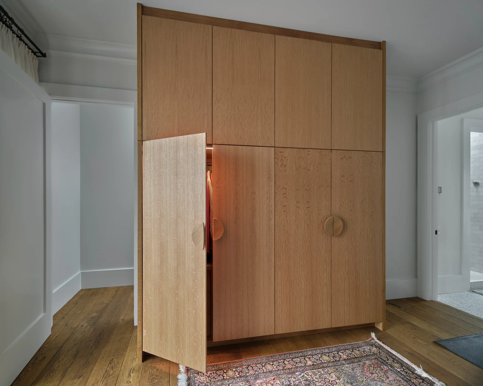 A wooden cabinet with its doors open in a room with white walls, hardwood floors, a patterned rug, and a partially visible door on the right.