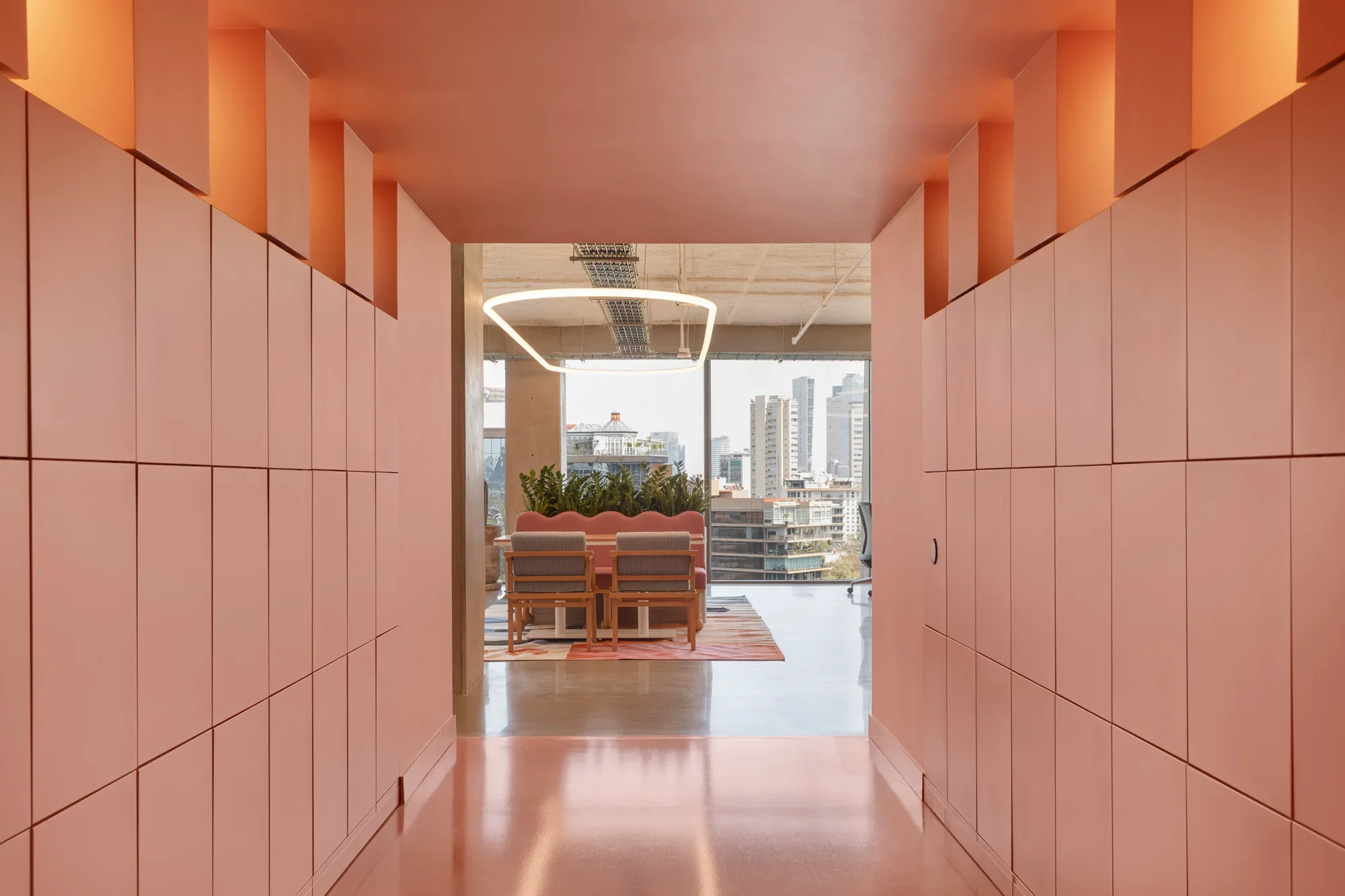 Pink hallway leading to a dining area with a round table and chairs, overlooking a city skyline through large windows, with a modern square light fixture hanging from the ceiling.