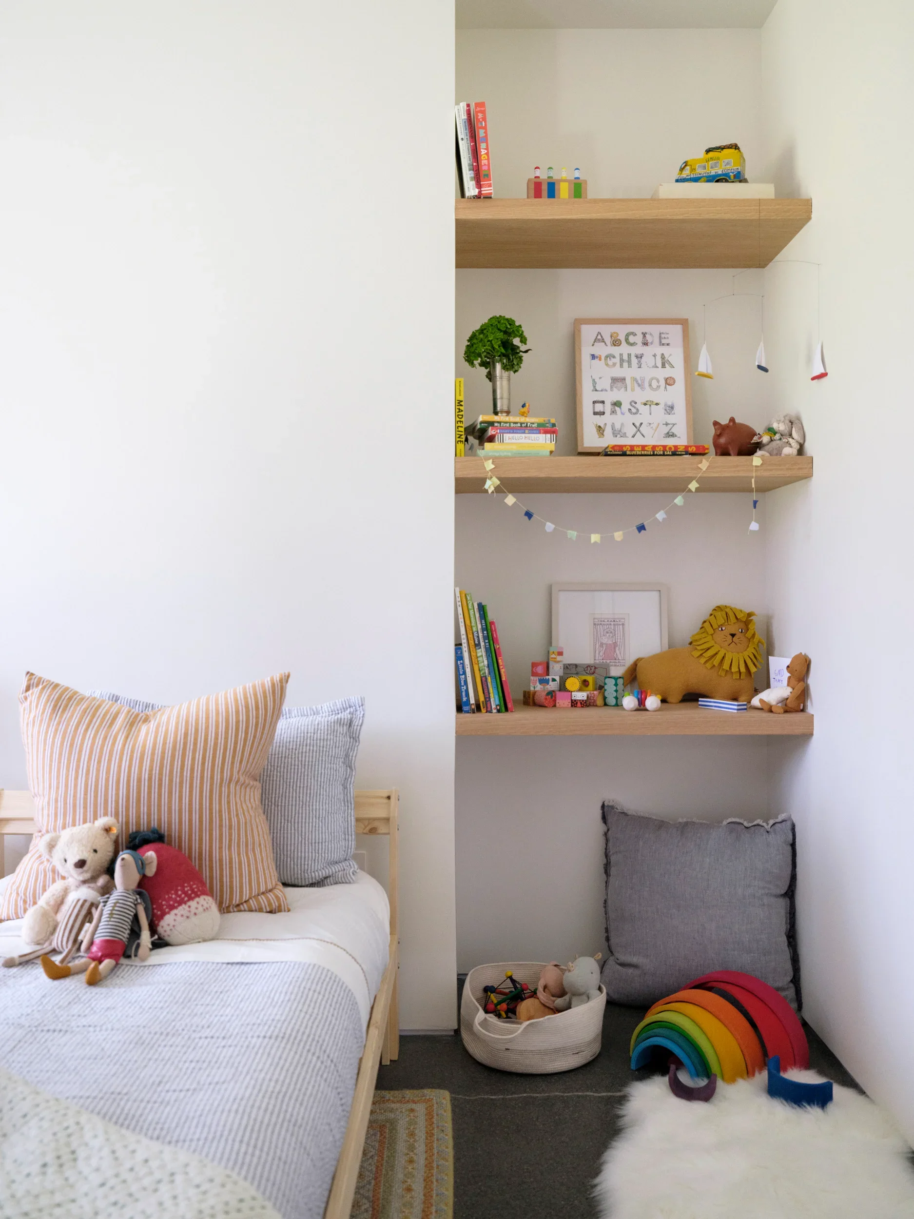 A cozy children's bedroom with a bed on the left and a wall with three wooden shelves on the right. The bed has brightly colored pillows and plush toys, including a teddy bear, a stuffed animal, and a sock monkey. The shelves hold books, toys, a framed alphabet print, and a small potted plant. The floor features a fluffy white rug and a basket with stuffed animals, as well as a rainbow-colored stack of cushions and some small toys.