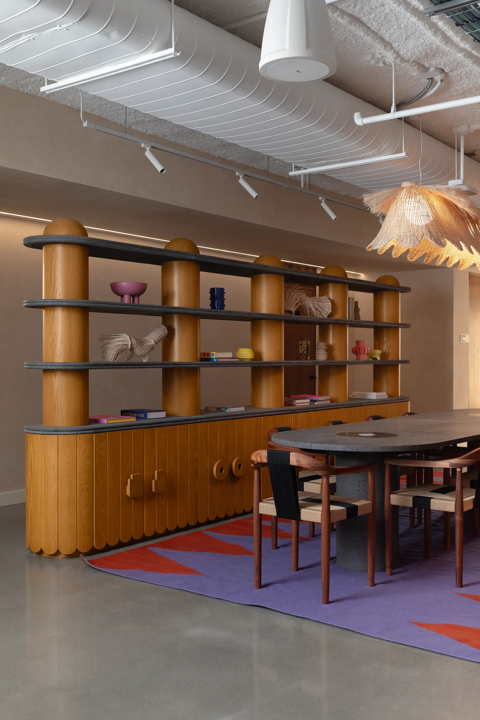 Interior shot of a modern room featuring a wooden shelf with curved edges, decorated with colorful vases and books. There is a round table with wooden chairs around it, a colorful patterned rug, and a unique, artistic pendant light hanging from the ceiling. The ceiling has exposed ducts, pipes, and track lighting.