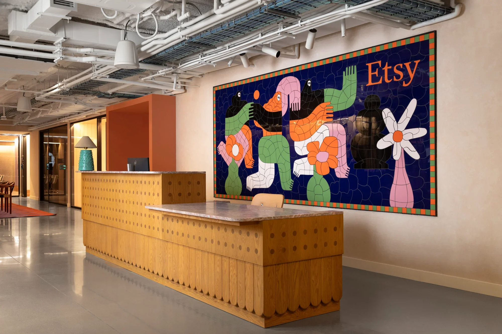 Interior of an office or lobby with a wooden reception desk, wall art featuring colorful abstract figures, and ceiling with exposed tubes, pipes, and lighting fixtures.