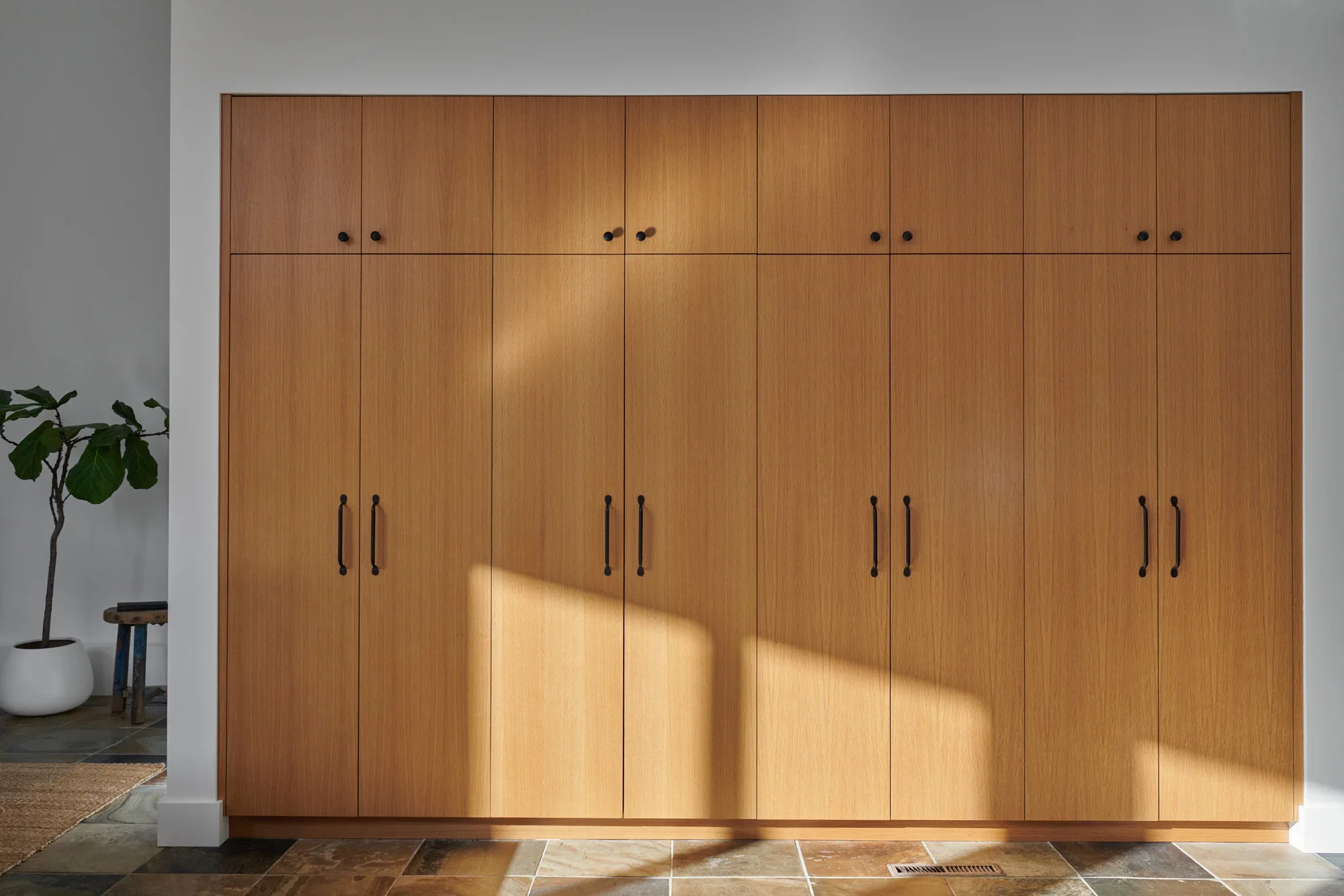 A wooden cabinet with black round handles, positioned against a white wall, with a potted plant on the left and tiled floor.