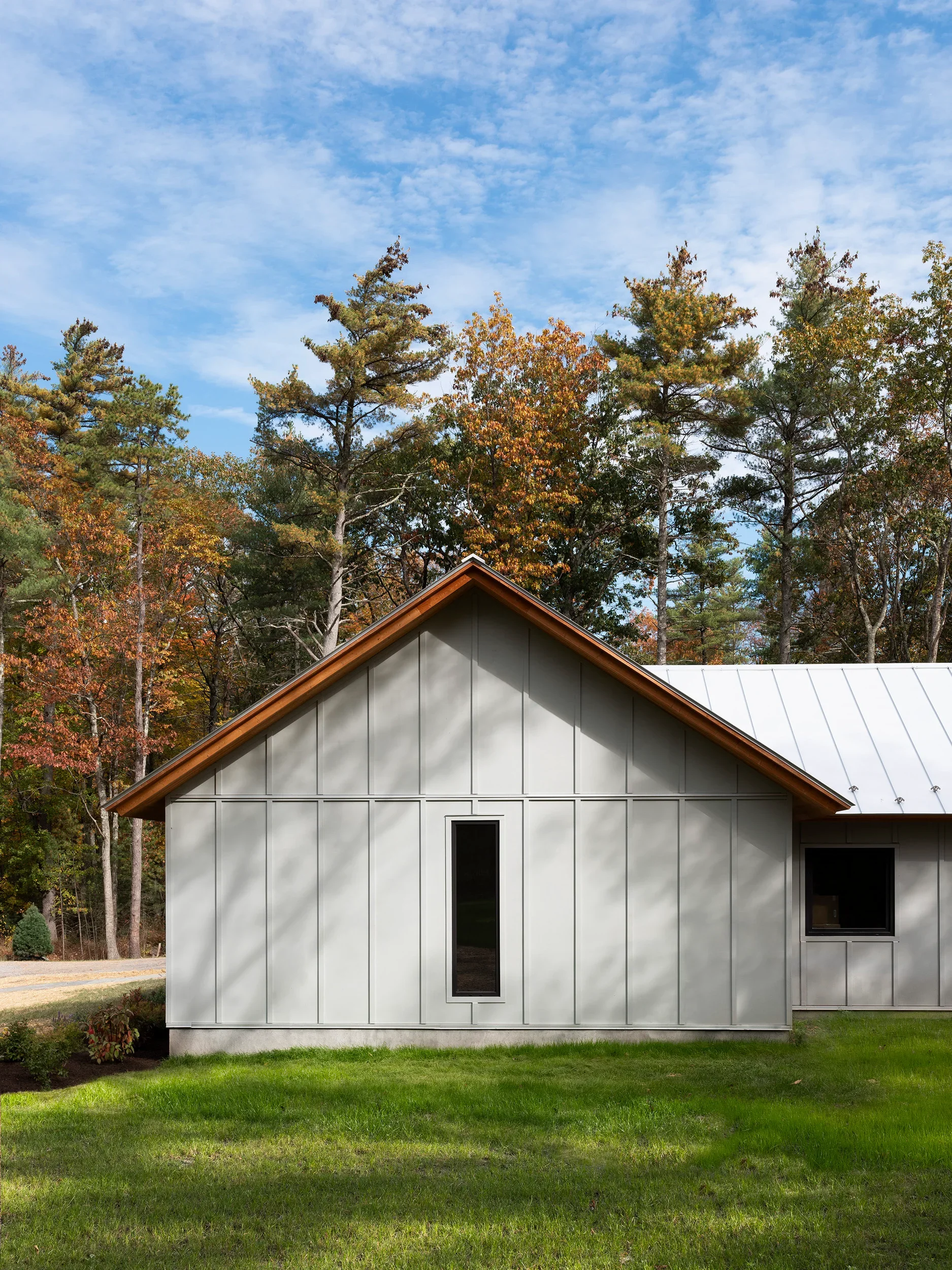 A modern house with gray exterior siding and a wooden roof edge, surrounded by green lawn and tall trees with autumn foliage, under a blue sky with scattered clouds.