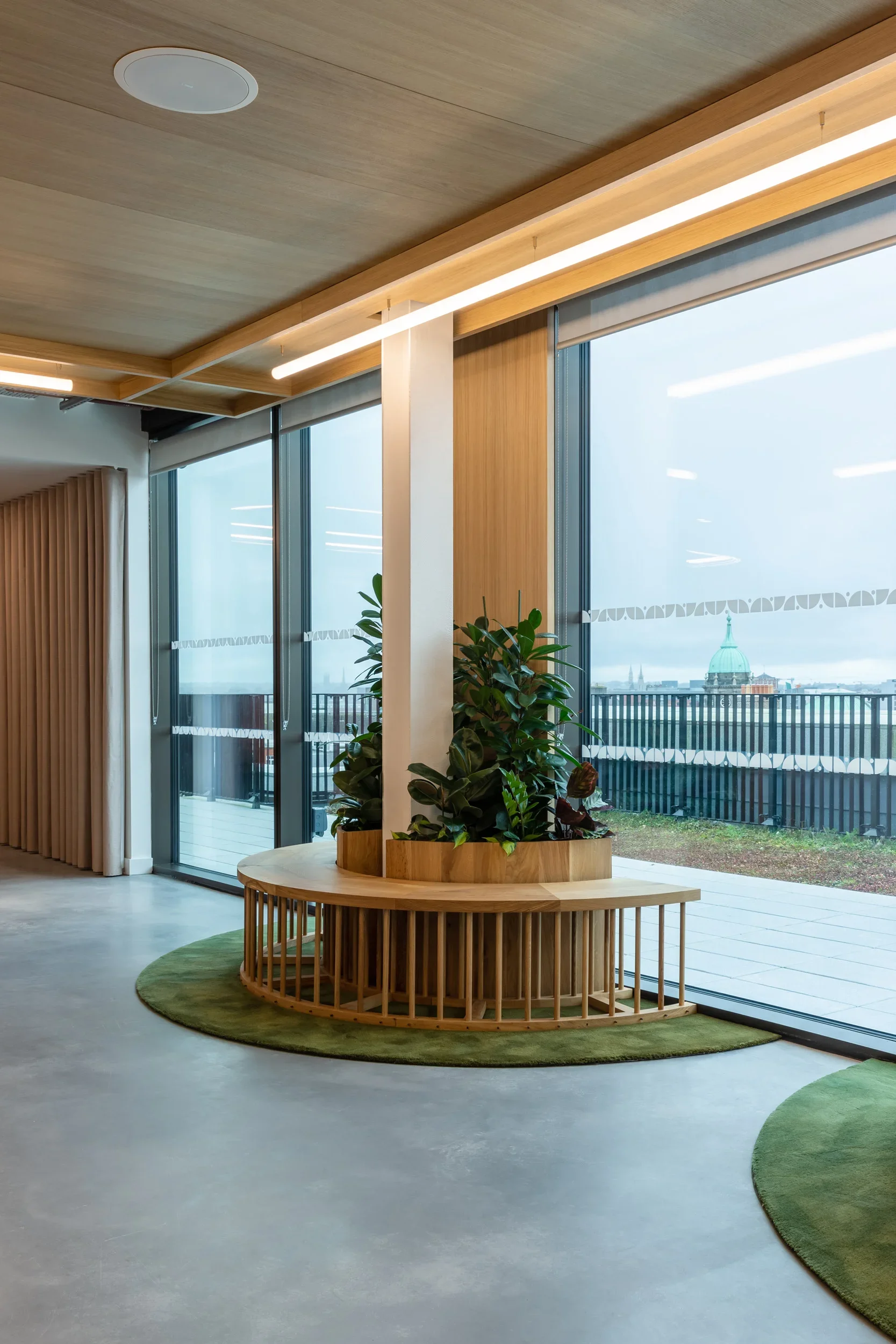 Interior view of a modern office or lobby with large floor-to-ceiling windows, wooden ceiling, plants, and a wooden circular planter with green plants. Outside the window, a cityscape with a prominent domed building is visible.