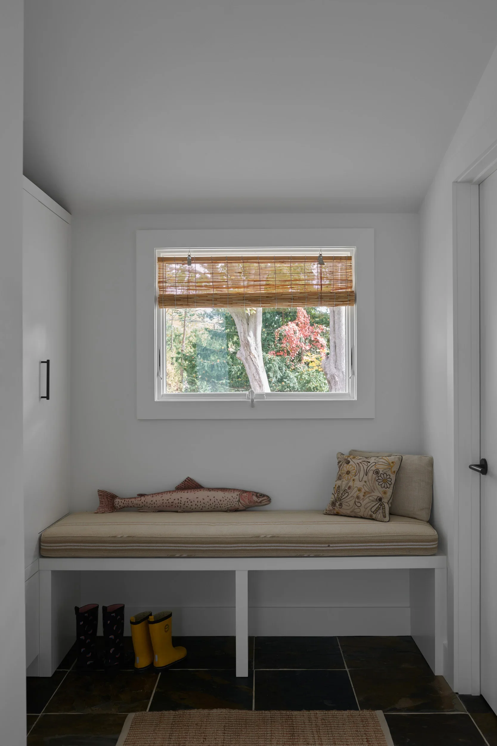 A built-in window seat with a striped cushion and pillows, a window with blinds overlooking trees, and multiple pairs of rain boots in a modern room.