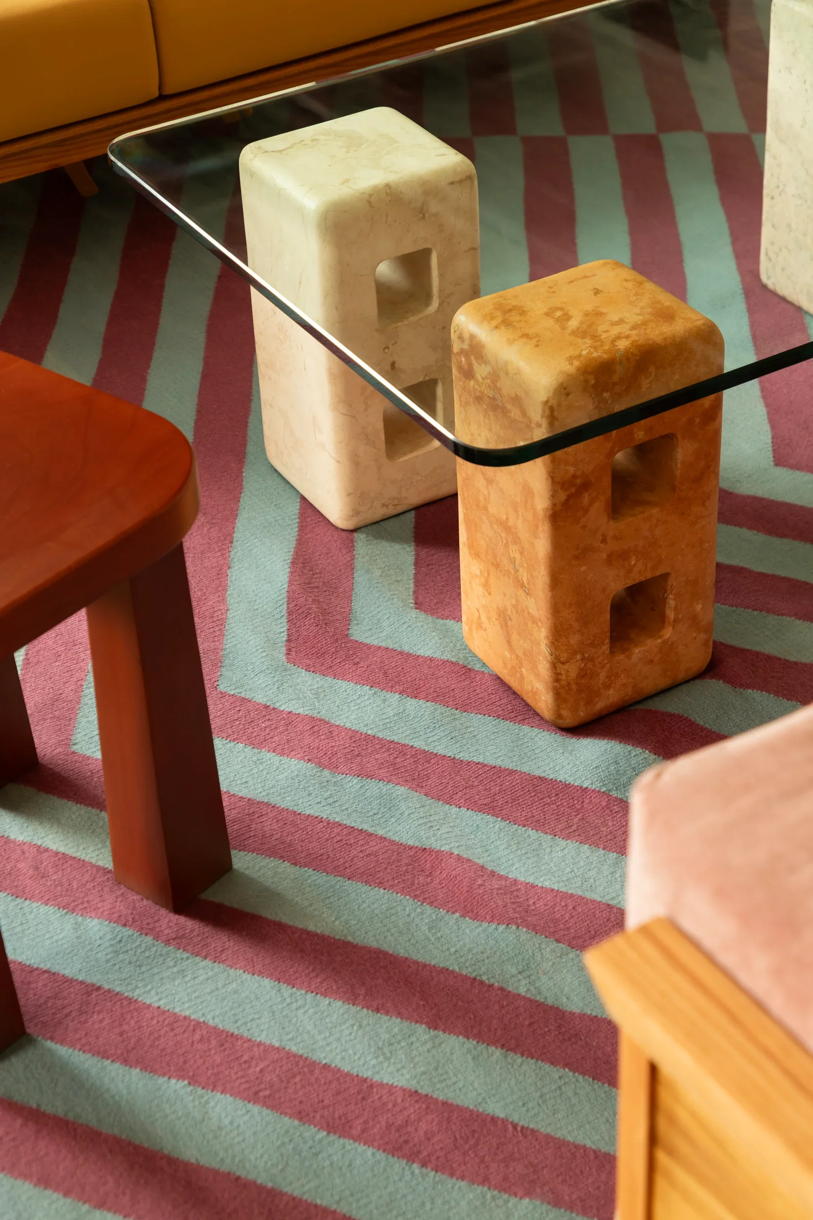 A glass table supported by two square stone or marble blocks, one beige and one orange, on a striped pink and green carpet. Part of a wooden chair and a brown couch are also visible.