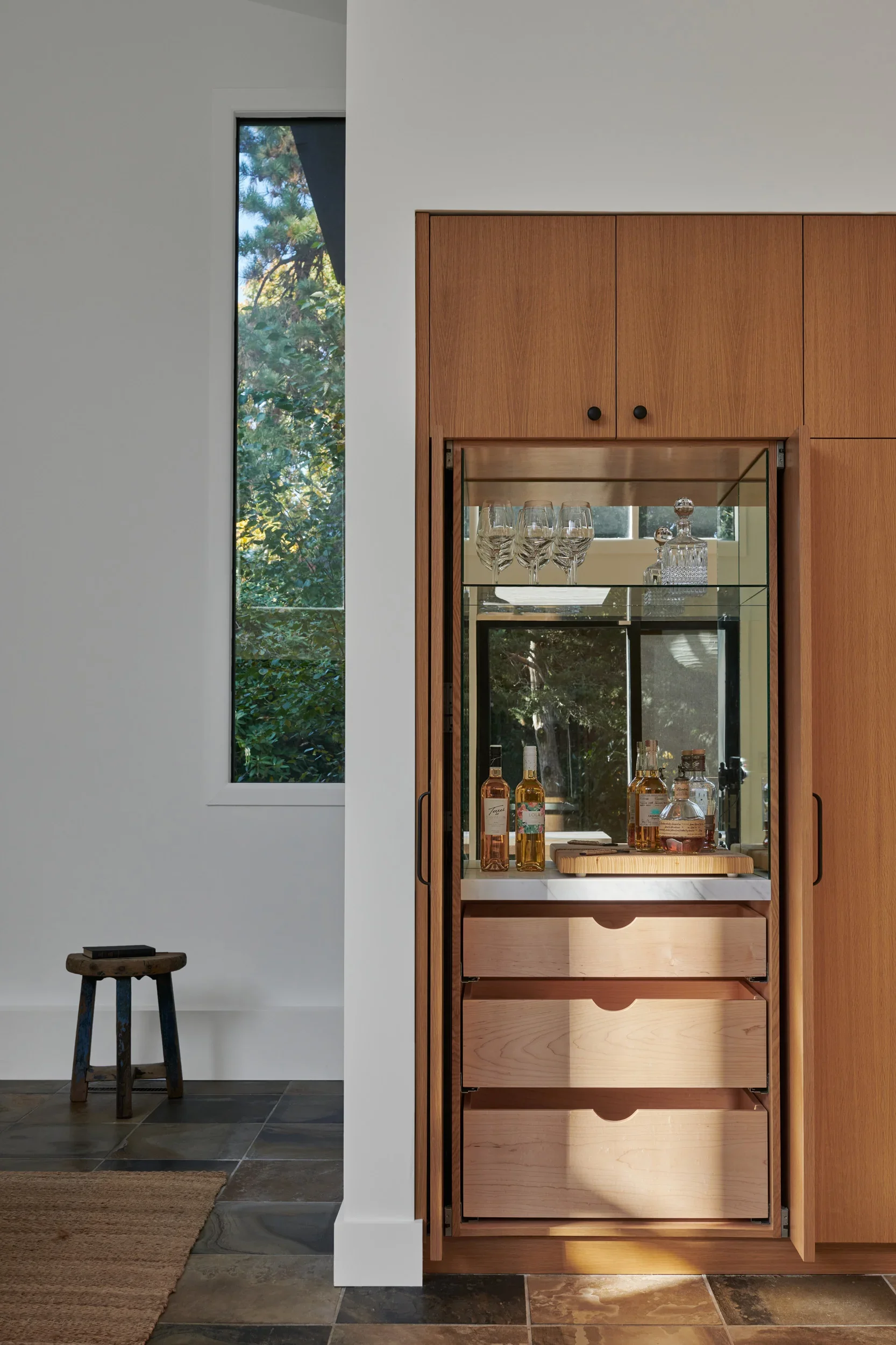 A wooden bar cabinet with glassware and bottles, situated in a modern room with a tall narrow window showing trees outside, and a small wooden stool on a tiled floor next to a white wall.