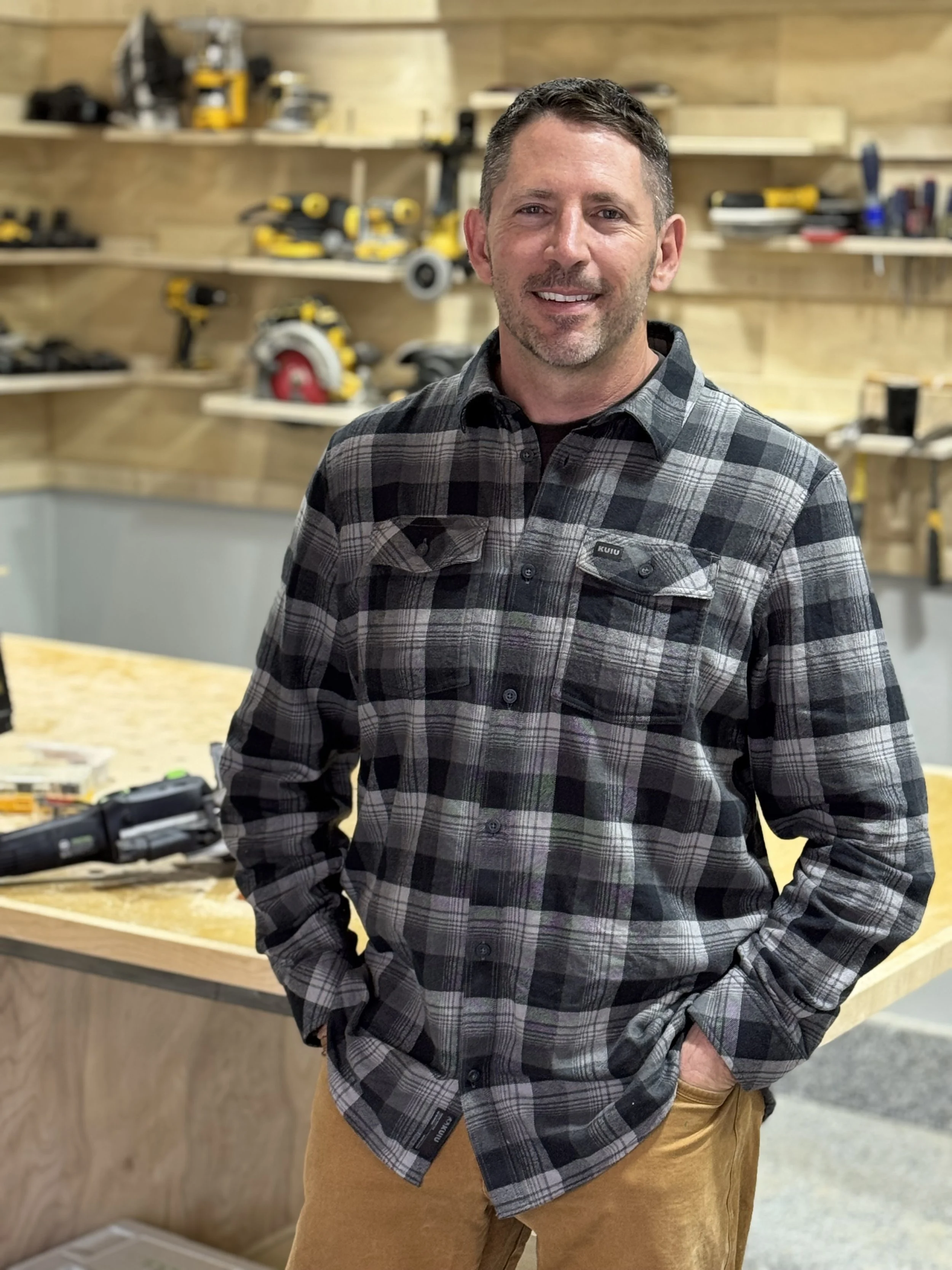 A man smiling, wearing a plaid shirt, standing in a workshop with tools on shelves behind him.