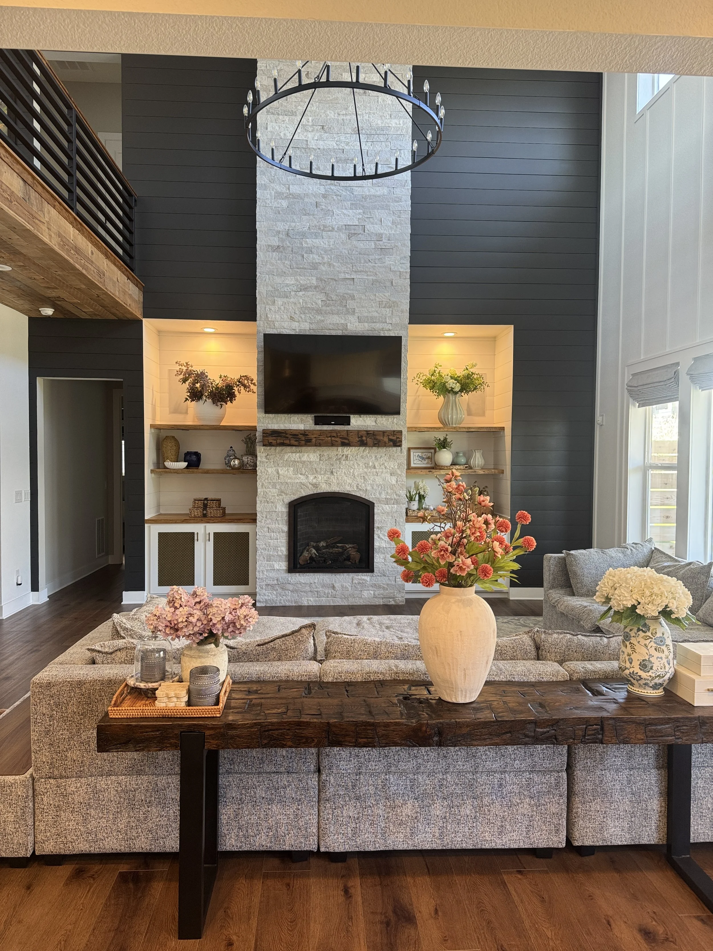 Living room with a large white stone fireplace, flat-screen TV above the fireplace, decorative shelves with vases and plants, a chandelier hanging from the ceiling, a beige couch with floral pillows, and fresh flowers in vases.