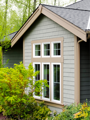 Side view of a house with a large rectangular window and shingle siding, surrounded by greenery.