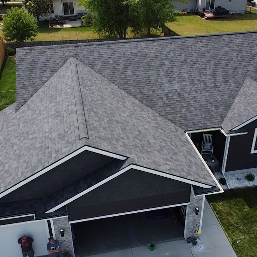 Aerial view of a residential house with a gray shingle roof, garage, and small porch, surrounded by grass and neighboring houses.