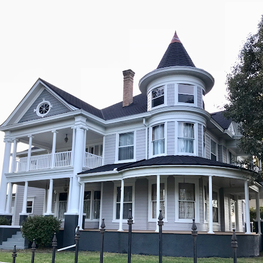 A large, white Victorian-style house with a turret, front porch, and multiple stories, surrounded by trees.
