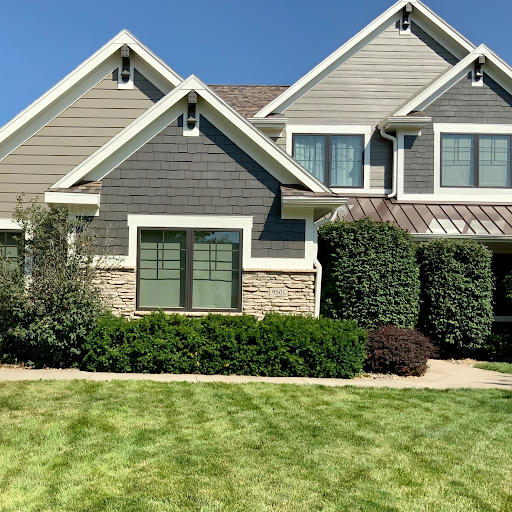 A modern suburban house with gray siding, large windows, and a well-maintained lawn with shrubs and trees in front.