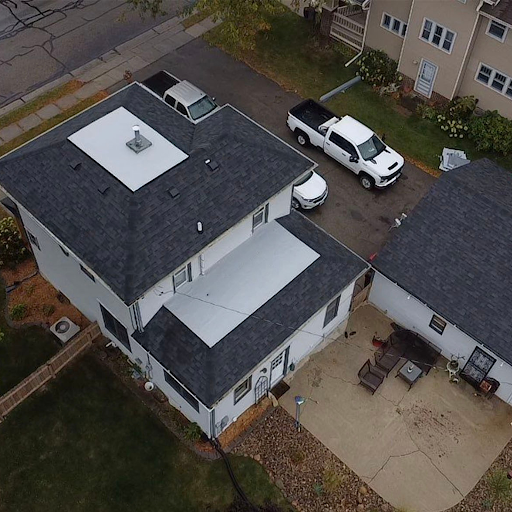 A two-story house with a dark roof, a small backyard, and a paved driveway with parked vehicles, including white and black trucks and SUVs, in a neighborhood with other houses.