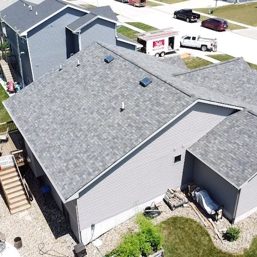 Aerial view of a residential house with a gray shingle roof, side yard, and neighboring houses in a suburban neighborhood.