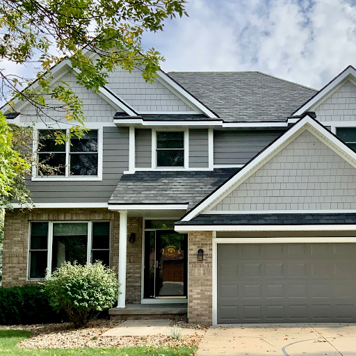 Front view of a two-story suburban house with a gabled roof, gray siding, brick accents, and a two-car garage. Green shrubbery and trees are in the yard.