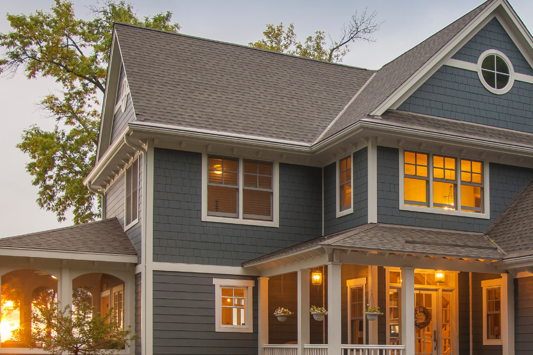 Two-story blue house with a covered front porch, multiple windows with warm lighting, and a gray roof, during sunset.