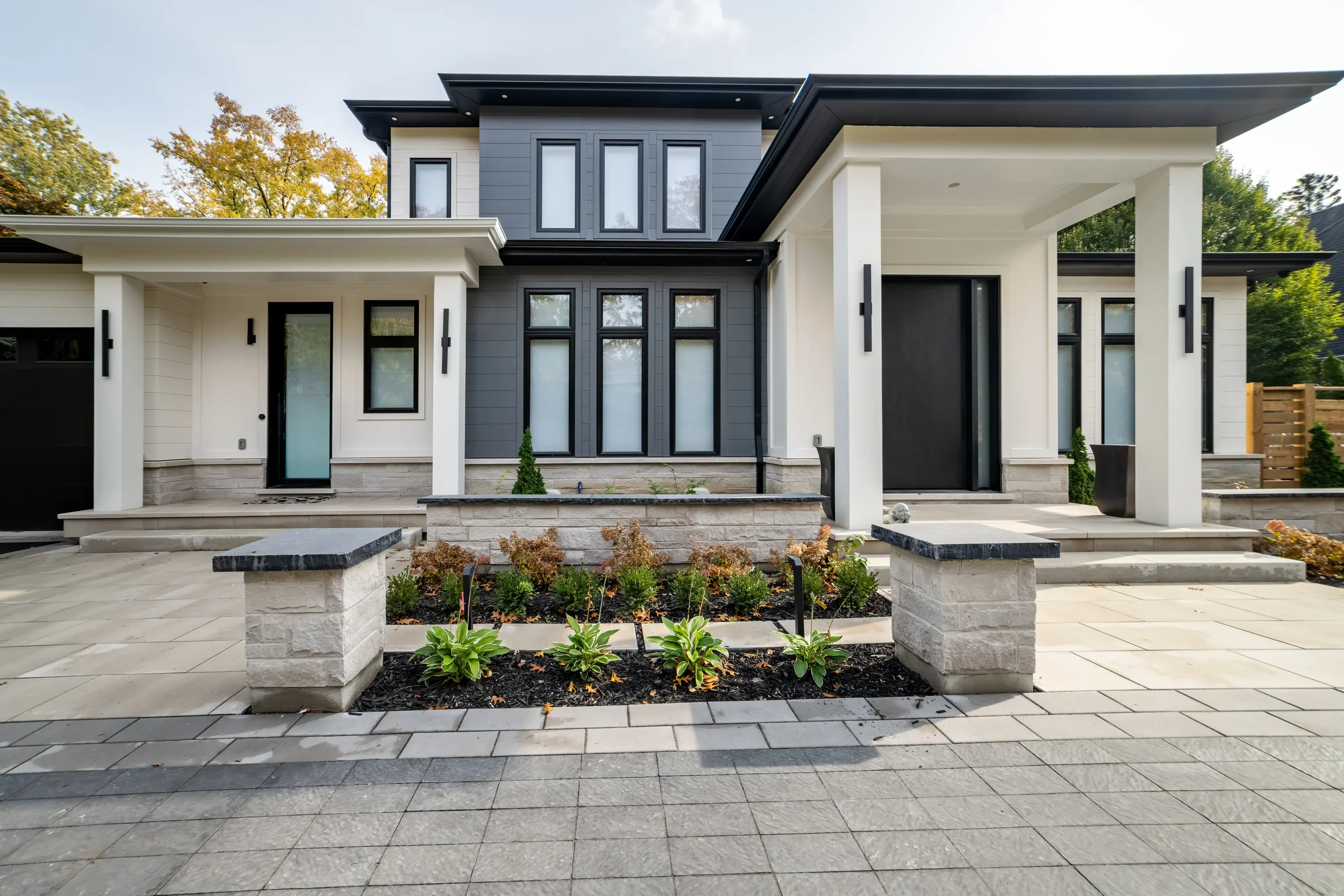 Modern two-story house with white and dark gray exterior, large windows, front porch, and landscaped yard with small plants and trees.
