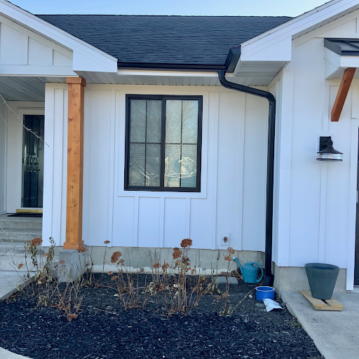 Front exterior of a house with a white wooden wall, window, black gutter, small garden bed, watering can, and potted plant.