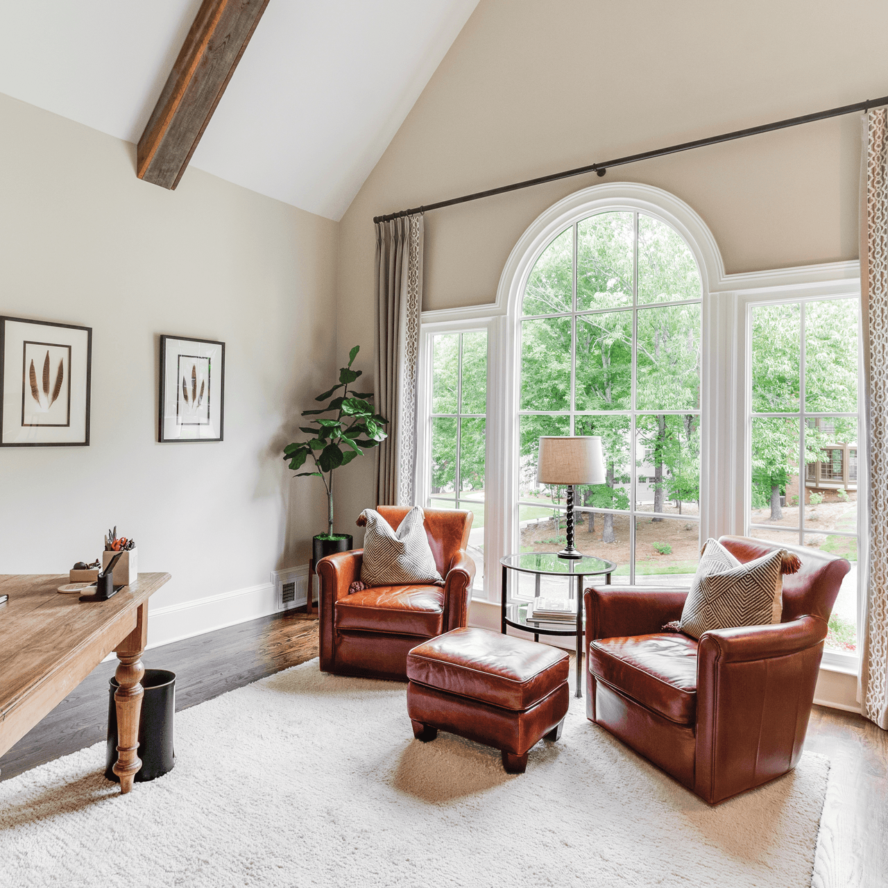 Living room with large arched window and two leather armchairs with pillows, a side table with a lamp, a potted plant, and decorative artwork on the wall.