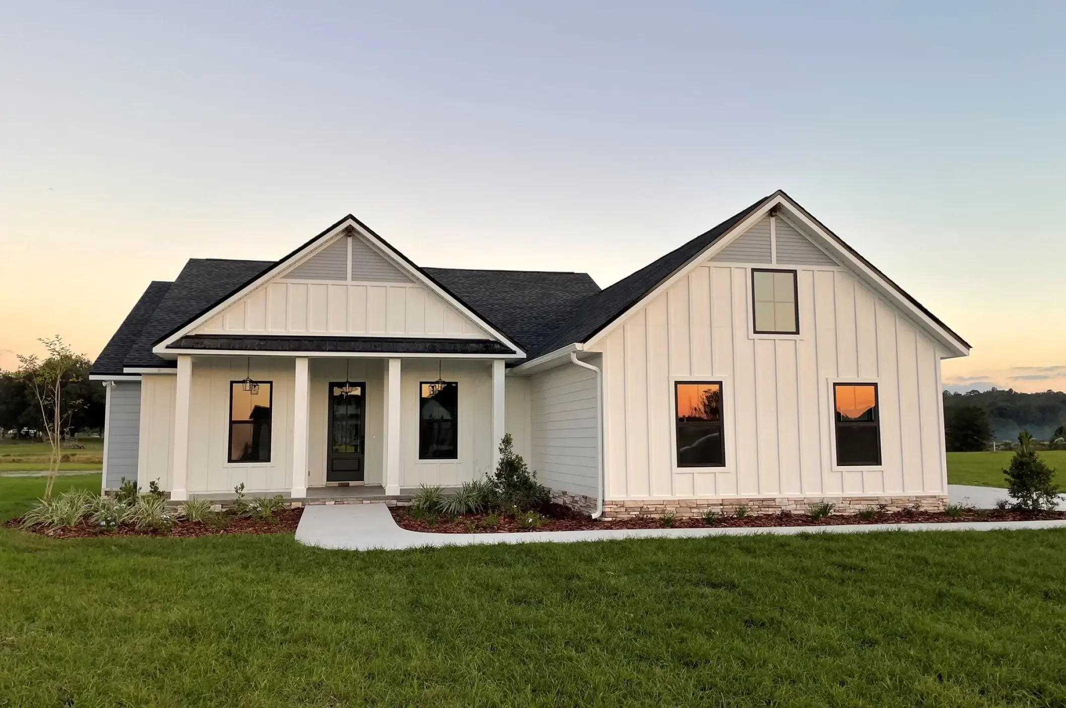 Front view of a modern farmhouse with white vertical siding, black roof, and large windows, set on a manicured lawn at sunset.