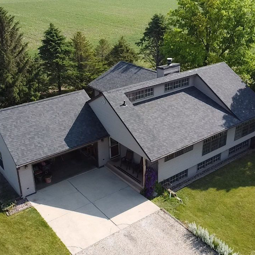 Aerial view of a two-story house with a gray roof, attached garage, a driveway, and a front porch, surrounded by green lawn and trees.