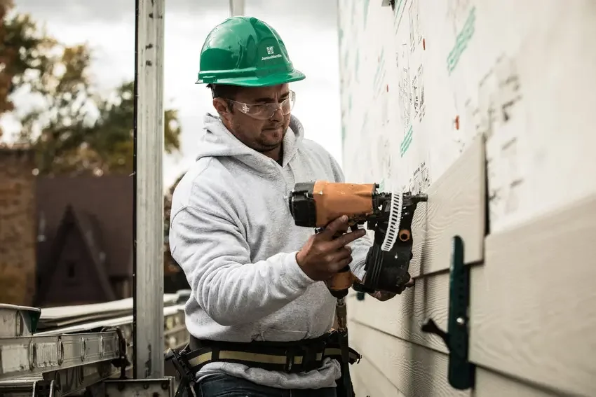 Man wearing a green hard hat and safety glasses using a nail gun on a house siding under construction.