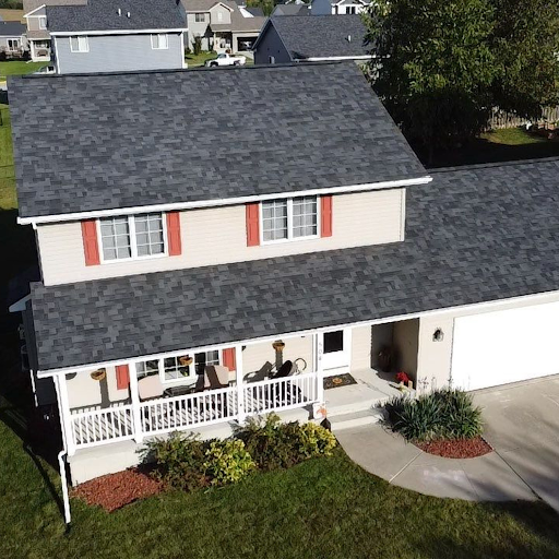 A two-story house with white siding, a black shingled roof, and red shutters on the windows. It has a front porch with white railings, decorated with potted plants, and a driveway leading to a garage. There are neighboring houses visible in the backg