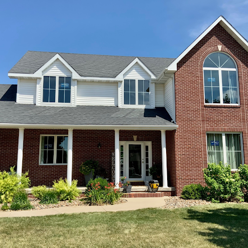 A large two-story house with a brick and white siding exterior, multiple windows, and a front porch with plants, in a well-kept lawn under a blue sky.