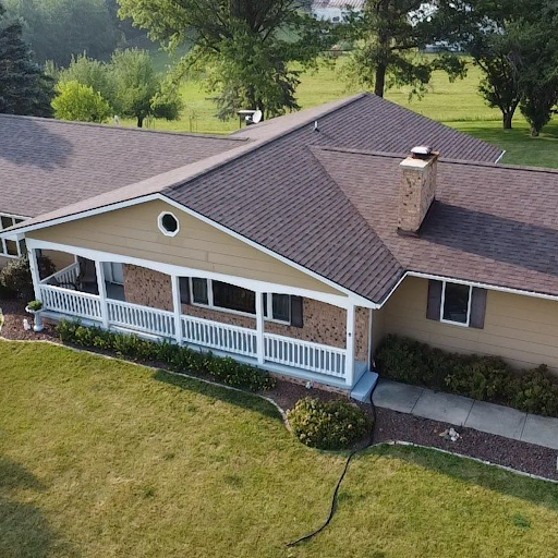 A house with a brown roof, beige siding, a brick chimney, and a white porch railing, surrounded by a lawn and trees.