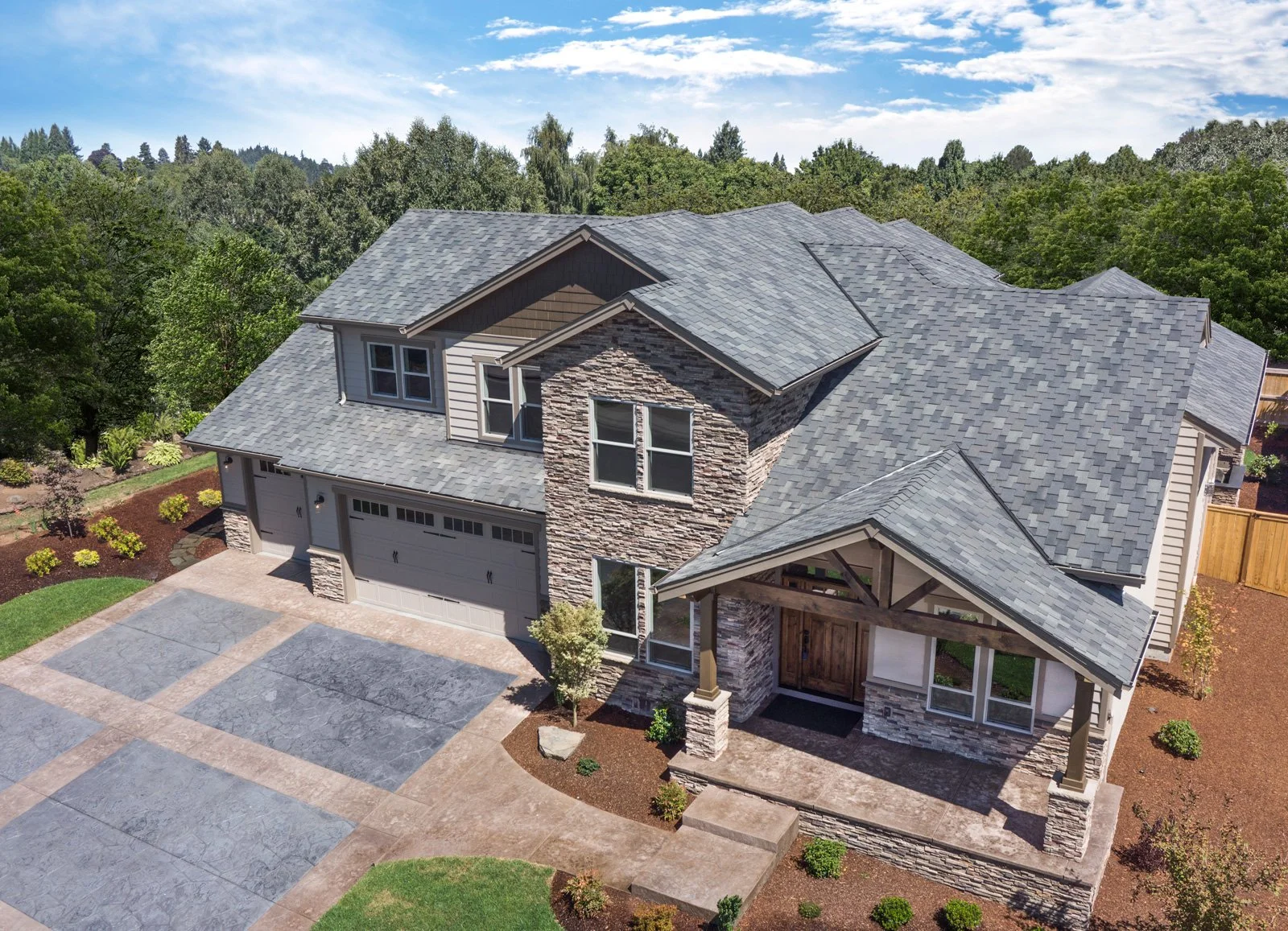 A two-story house with a gray shingle roof, stone accents, and a front porch, surrounded by a landscaped yard and trees.