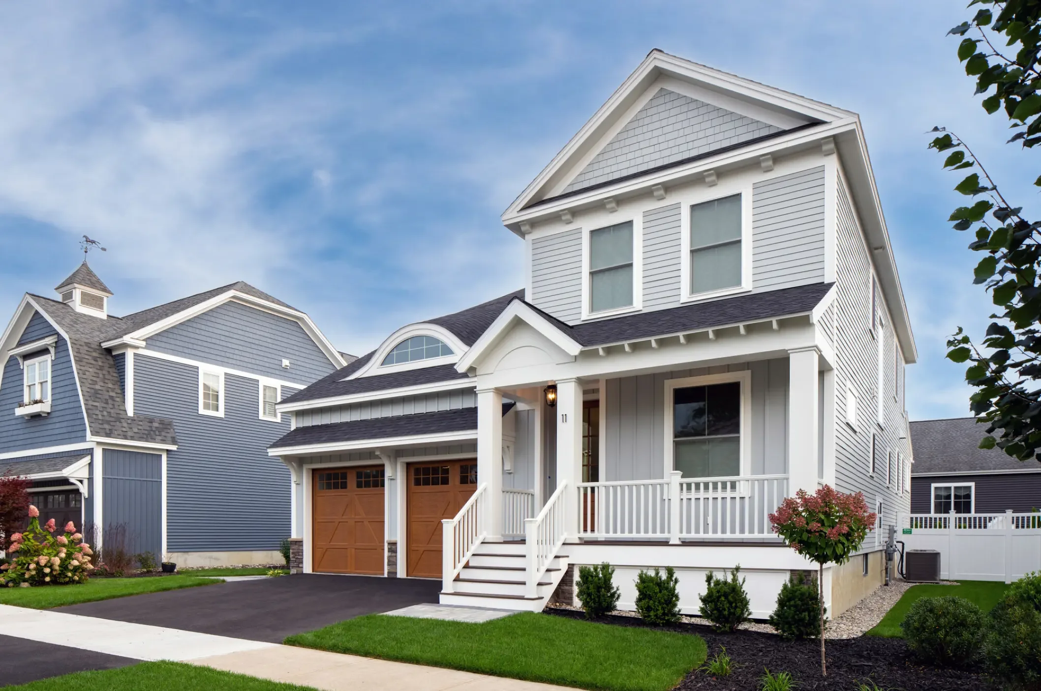 Front view of a modern two-story house with a grey and white exterior, a front porch with stairs, a wooden garage door, a driveway, a well-maintained lawn, and neighboring houses with similar architectural styles, under a partly cloudy sky.
