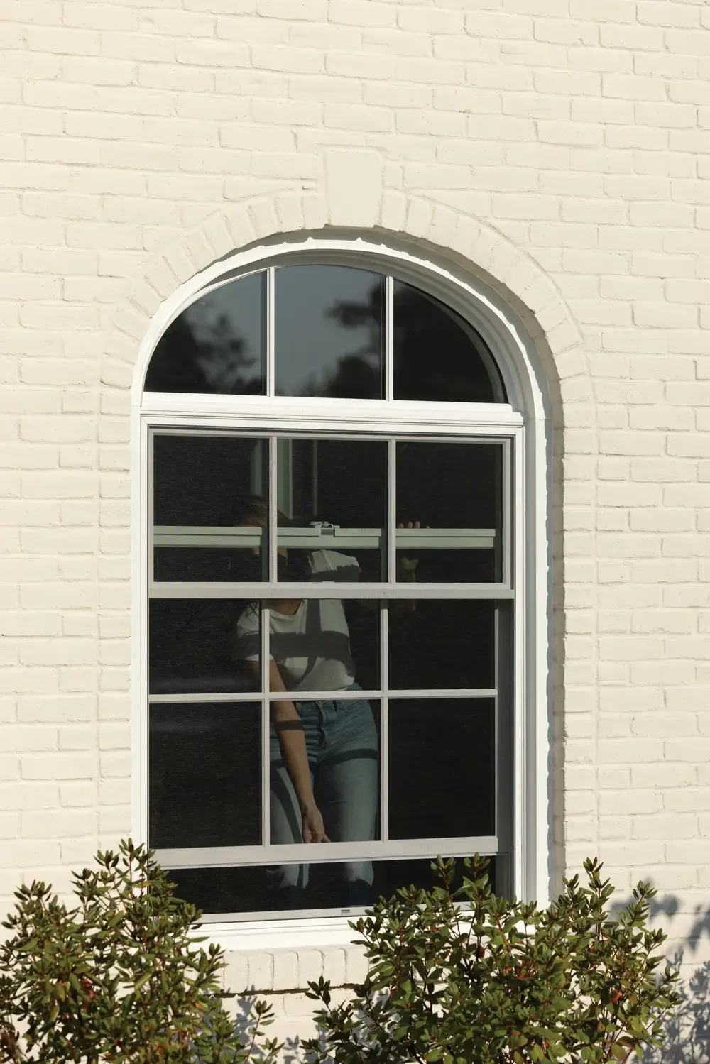 A white brick house with an arched window reflecting the sky and trees, with a person in casual clothing visible through the glass.